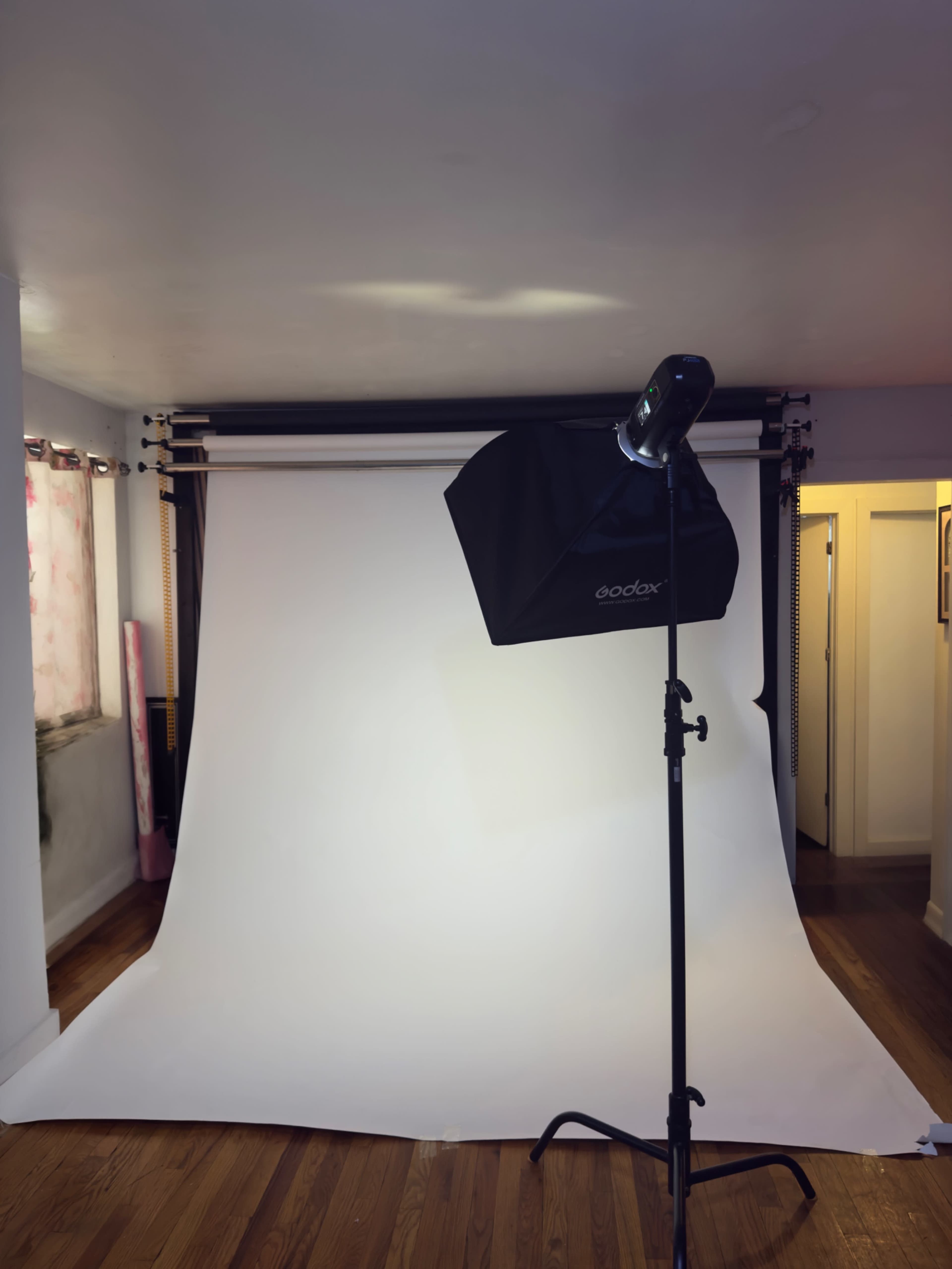 The image shows a photography studio setup featuring a white backdrop, a light stand with a softbox, and hardwood flooring.