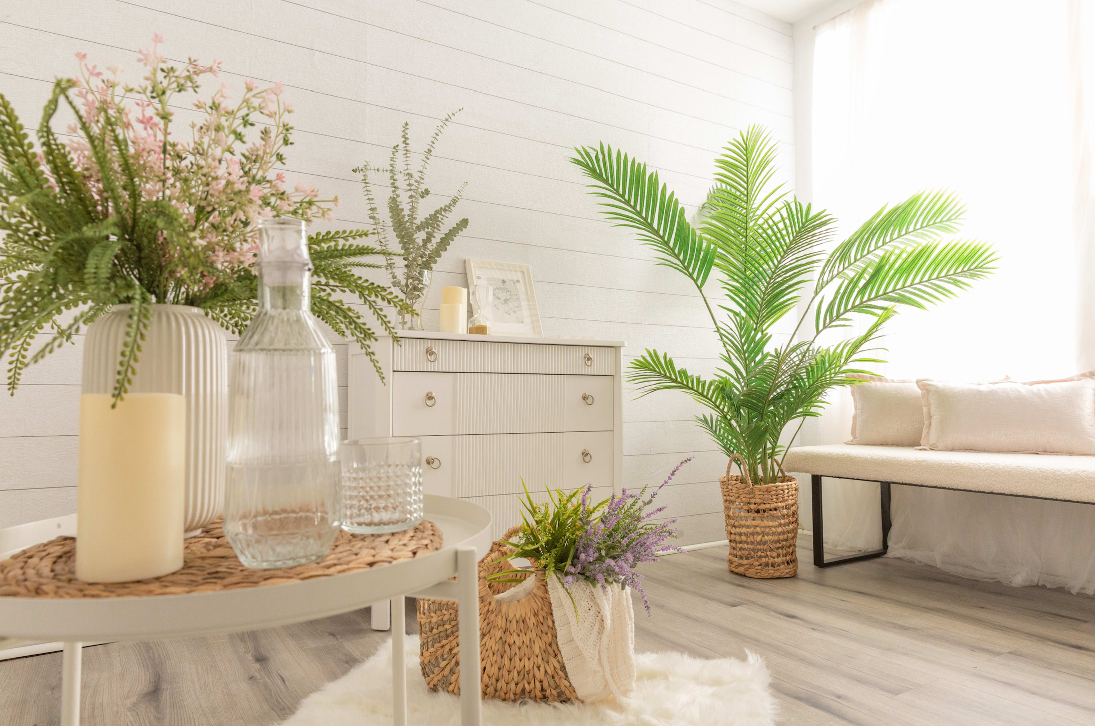 A cozy interior scene features a white dresser adorned with decorative plants, a glass water bottle, candles, and woven baskets on a light wood floor.