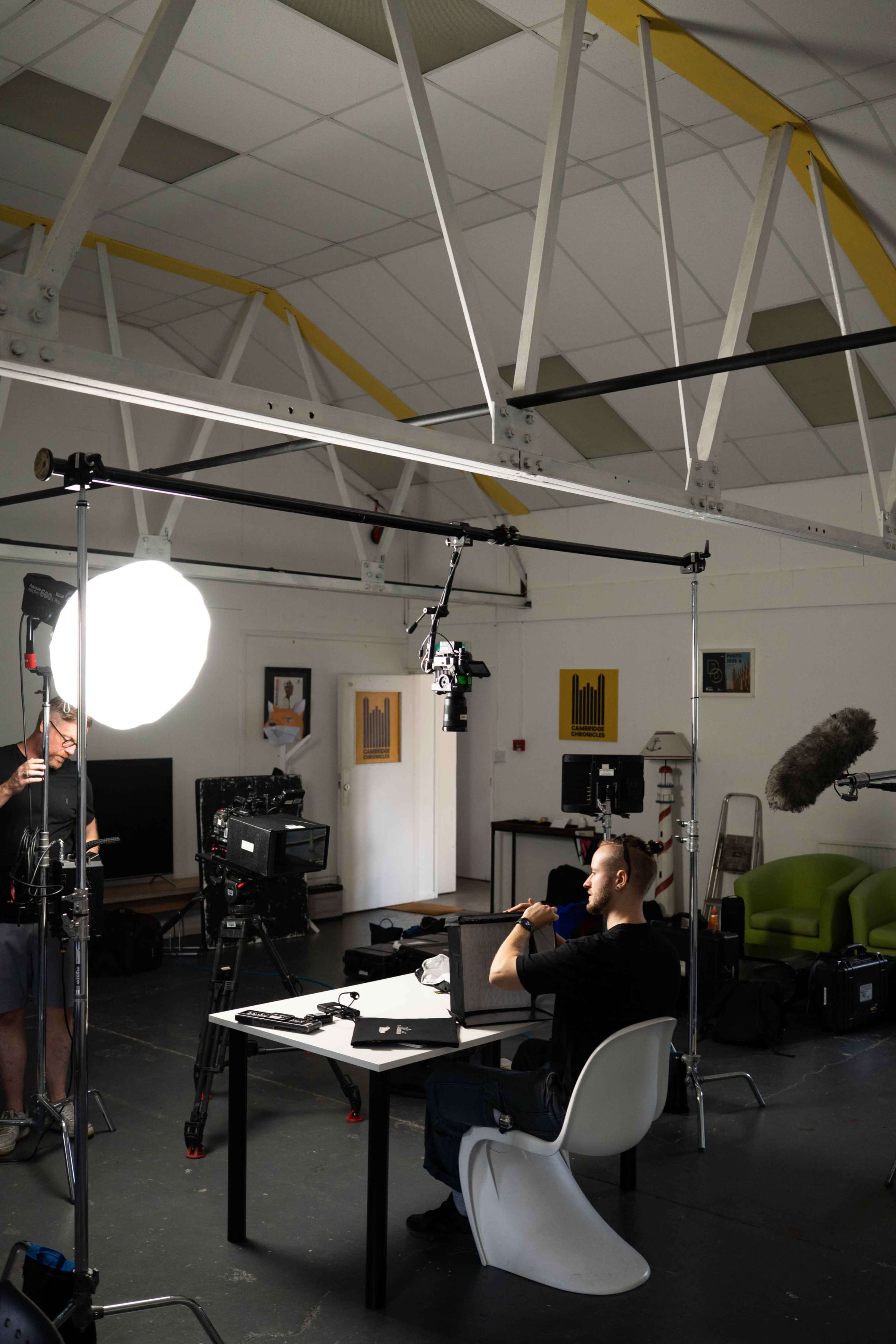 A man sits at a table in a well-lit studio while a crew sets up cameras and lights around him.