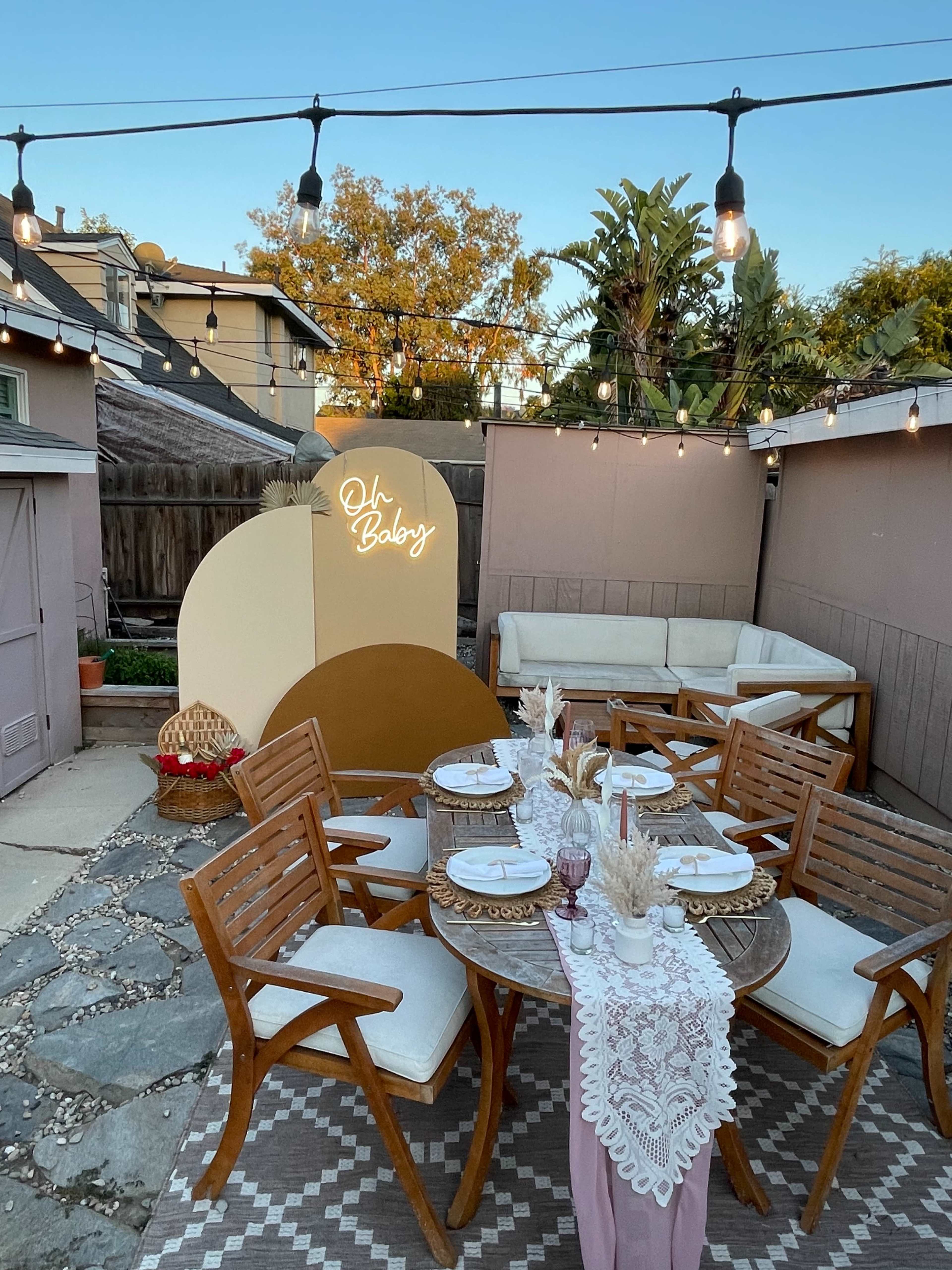 A cozy outdoor seating area features a round table set with chairs, decorated with a lace tablecloth and surrounded by warm string lights.
