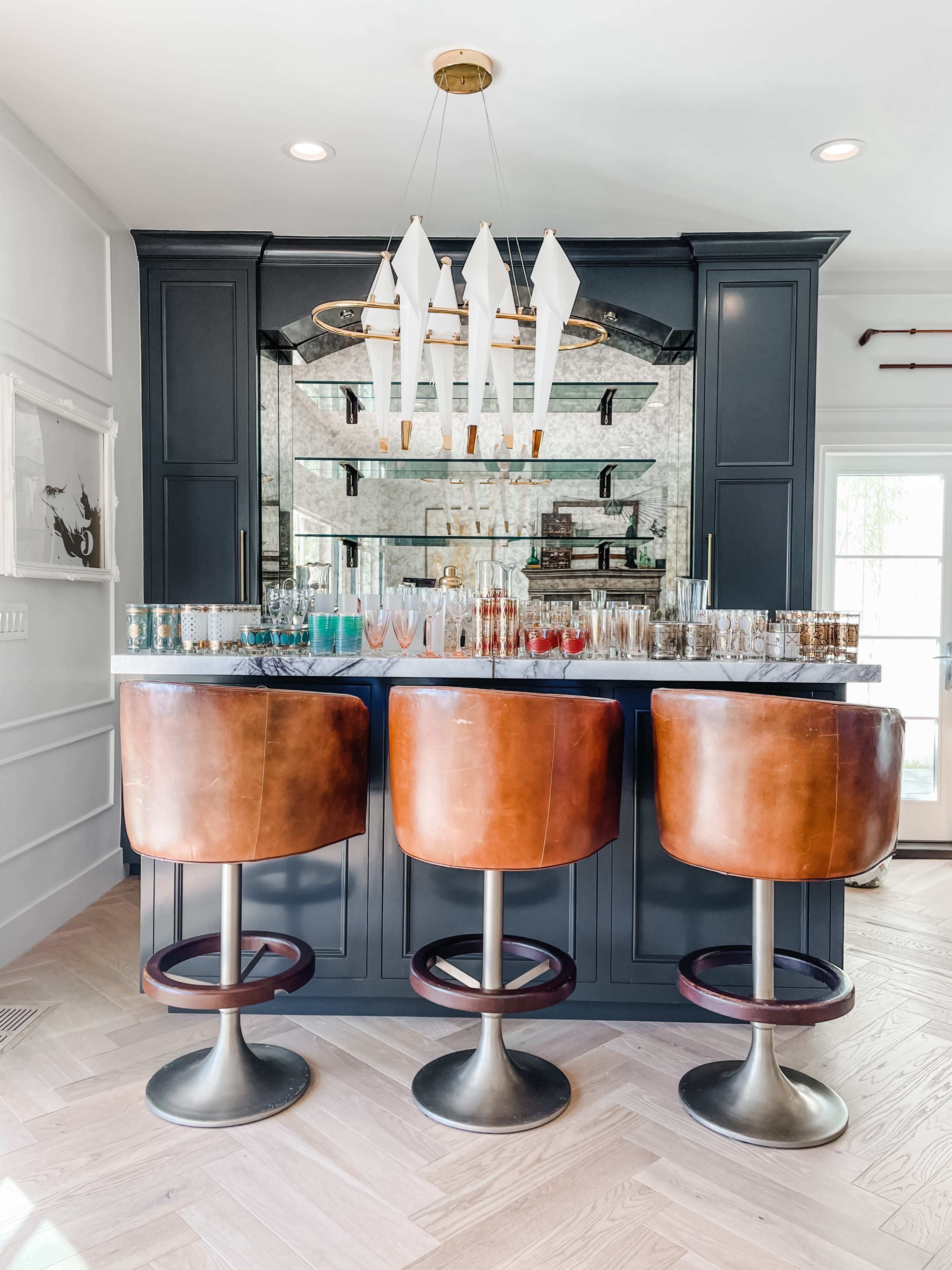 A modern bar area features three brown leather stools in front of a mirrored backsplash and a variety of colorful glassware on the countertop.