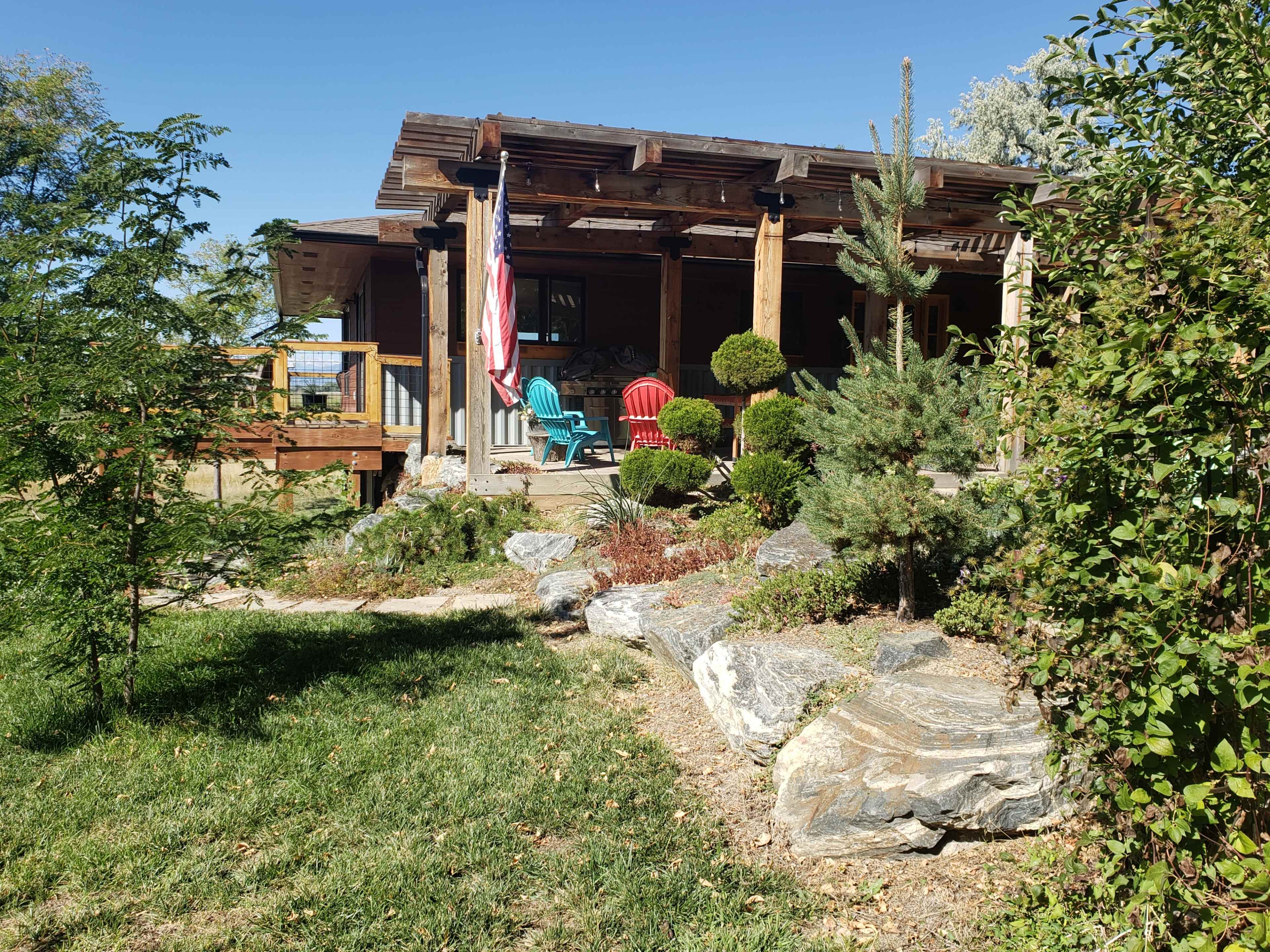 A wooden house with a porch features red and teal chairs, an American flag, and landscaped greenery with rocks and shrubs.