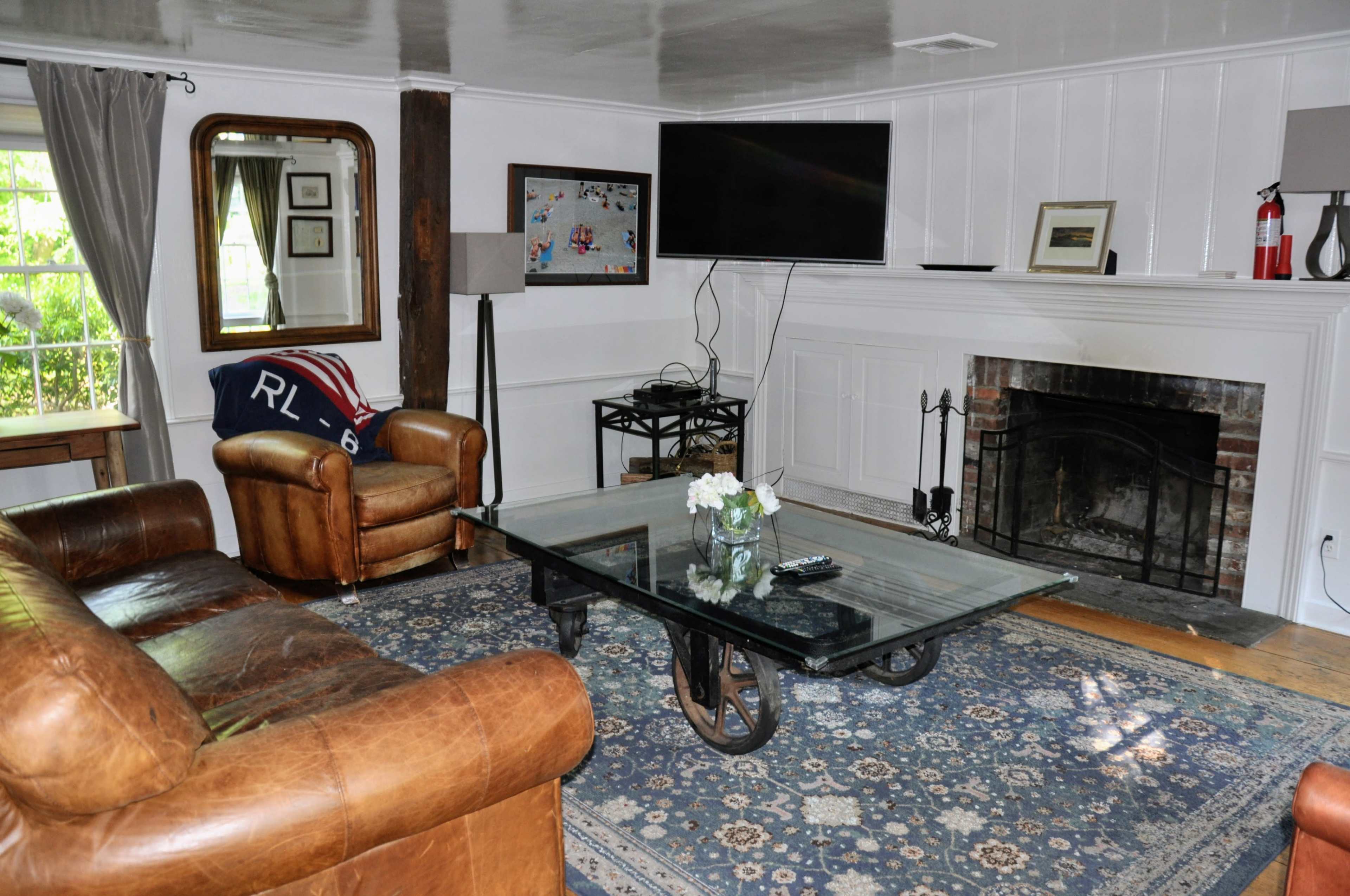 The living room features a leather chair, a glass coffee table on a patterned rug, and a television mounted above a white brick fireplace.