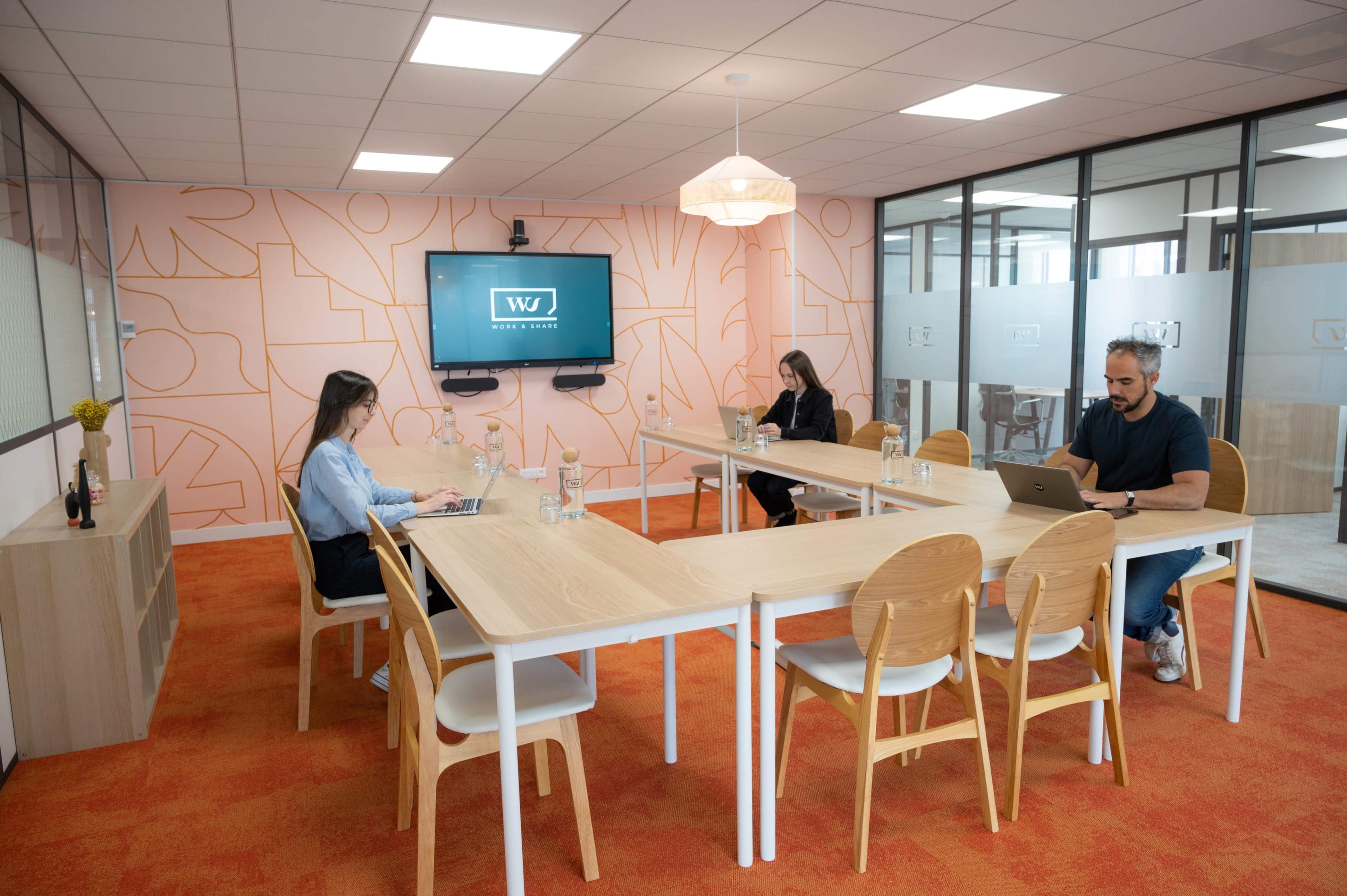 A modern conference room features three individuals seated at a table, with a large screen displaying a logo on the wall.