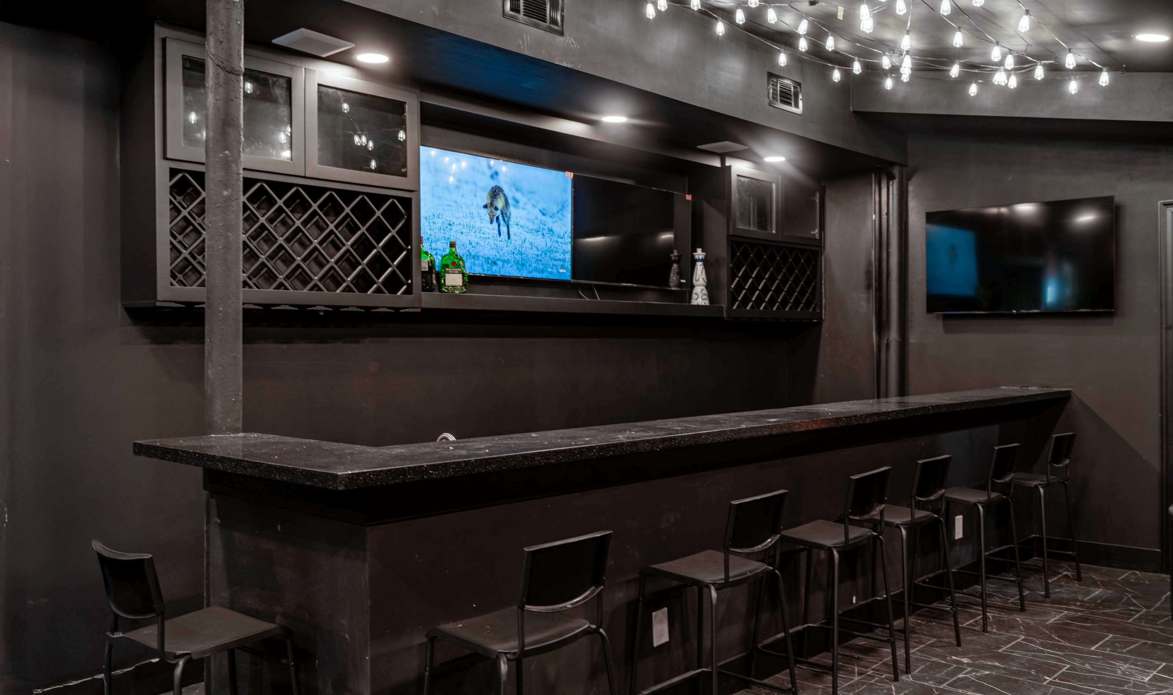The image shows a dimly lit bar area with a long black countertop, several high stools, and multiple screens mounted on the wall.