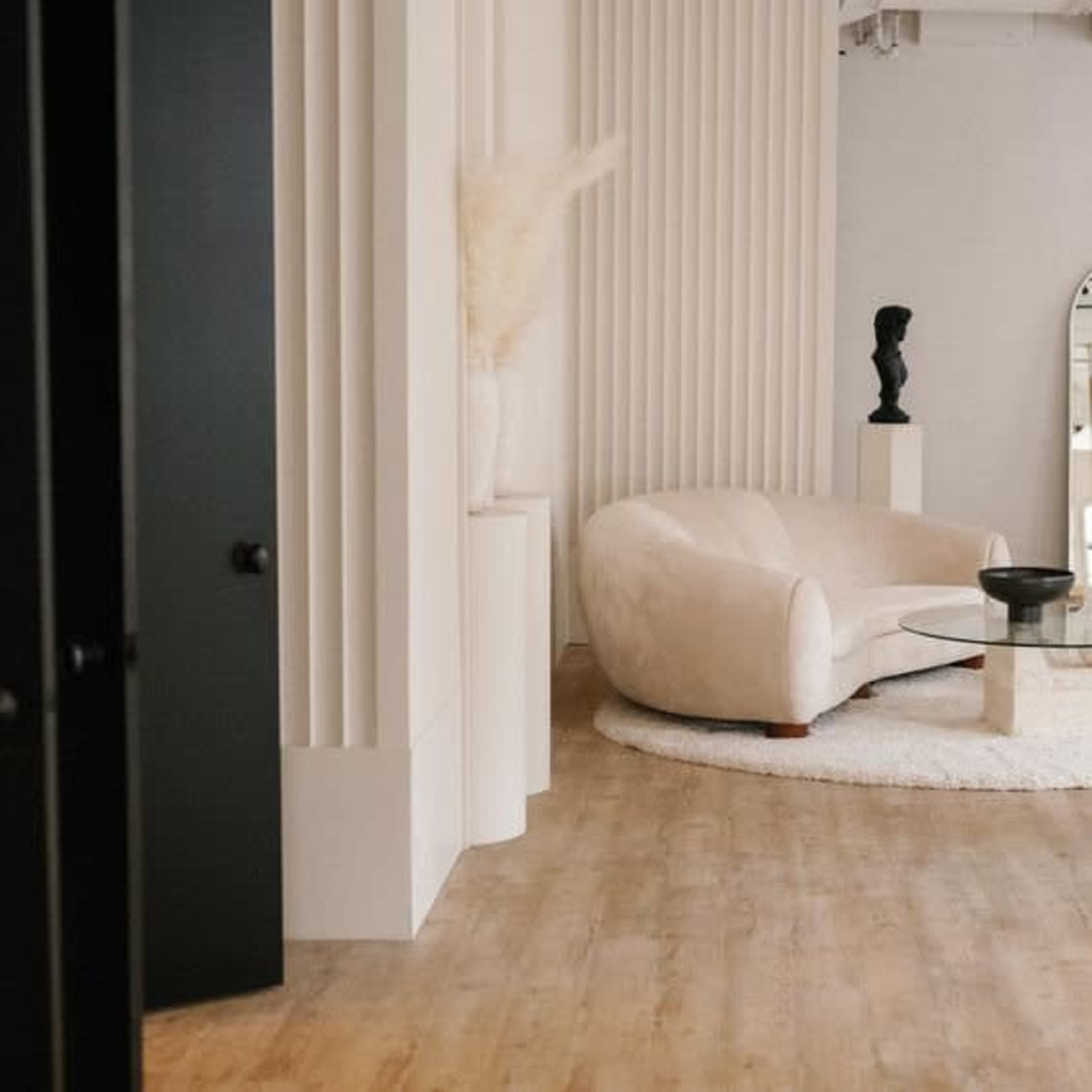 A modern interior featuring a curved beige sofa, a round glass coffee table, and minimalist decor against textured white walls.