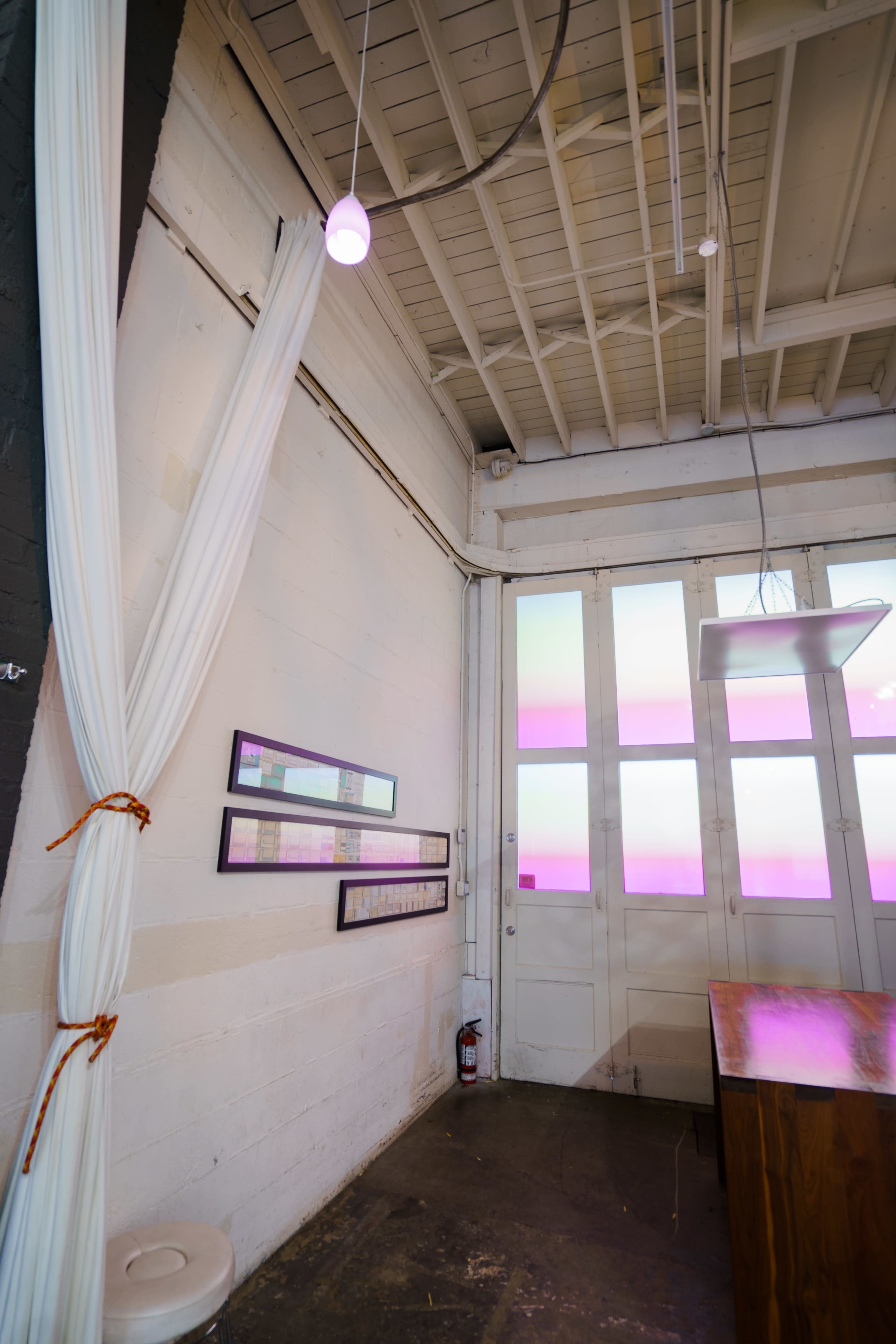 An interior space with white walls, curtains, a wooden table, framed artwork, and a large window with pink-tinted glass.