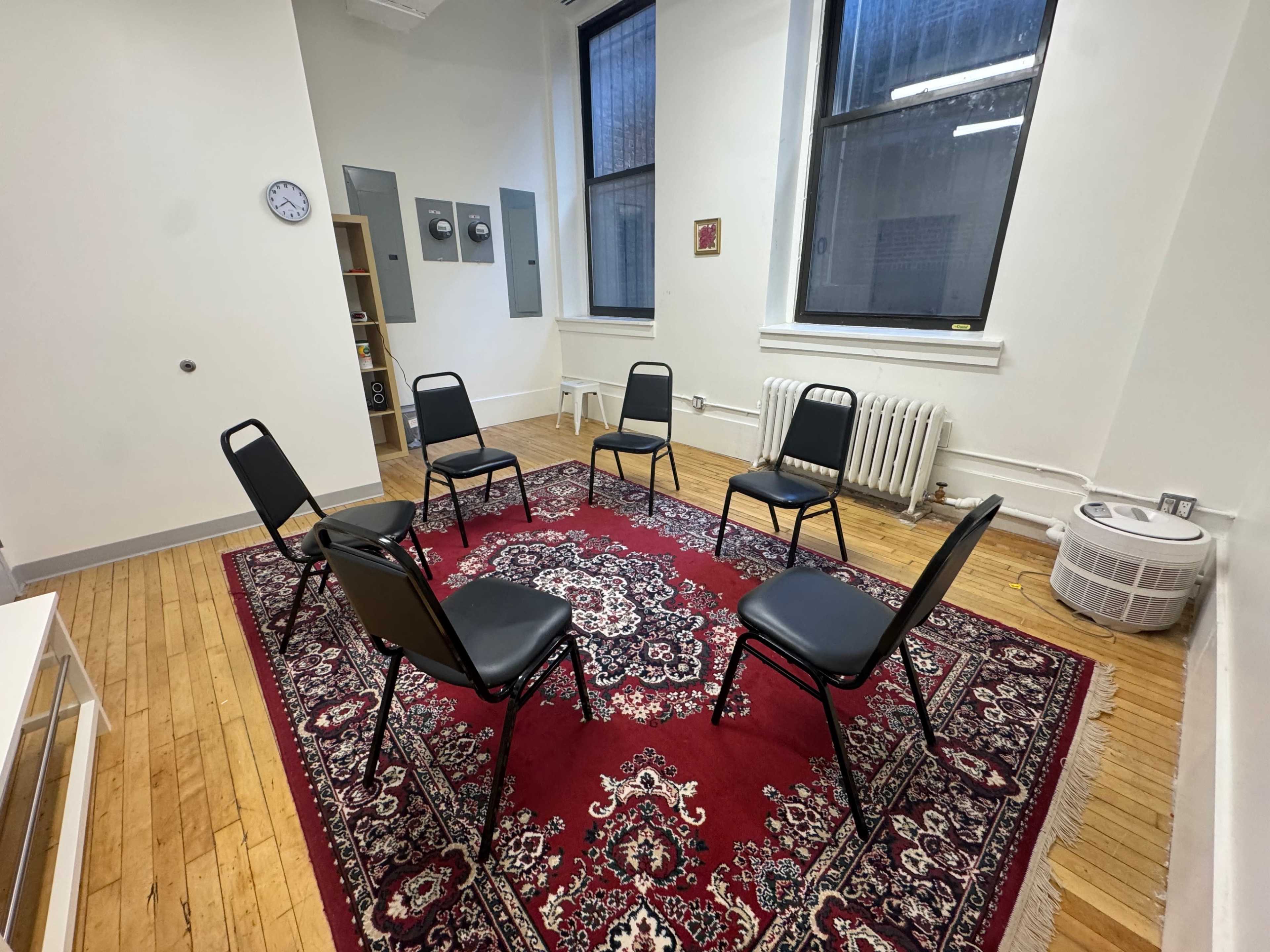 The image shows a room with six black chairs arranged in a circle on a red patterned rug, with a clock and electrical panels on the walls.