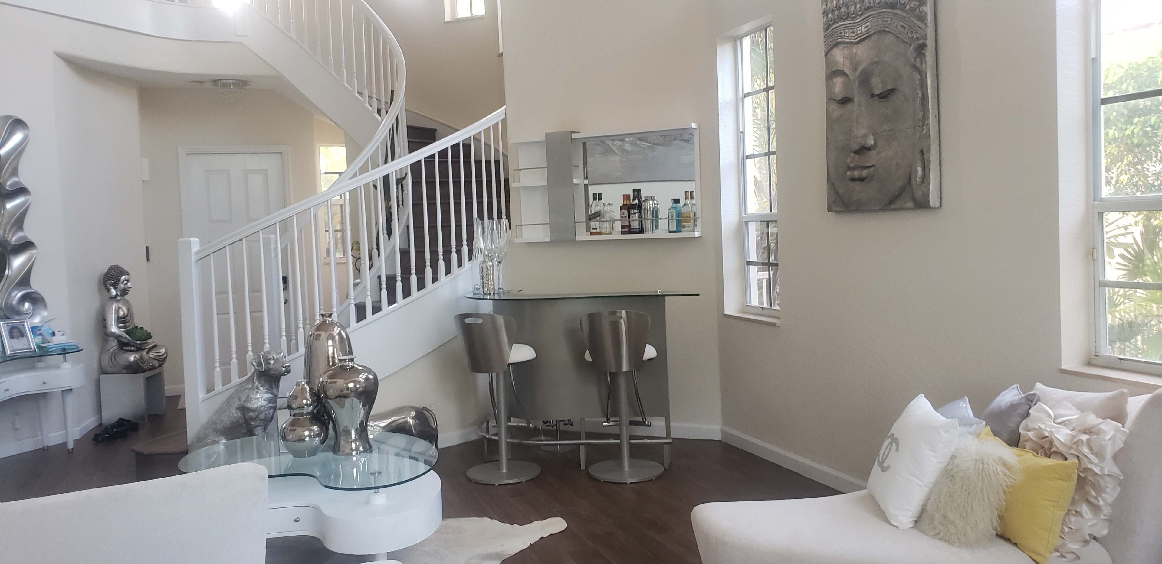 A spacious, modern living area featuring a curved staircase, a glass bar with stools, and various decorative elements, including a Buddha wall art and silver accents.