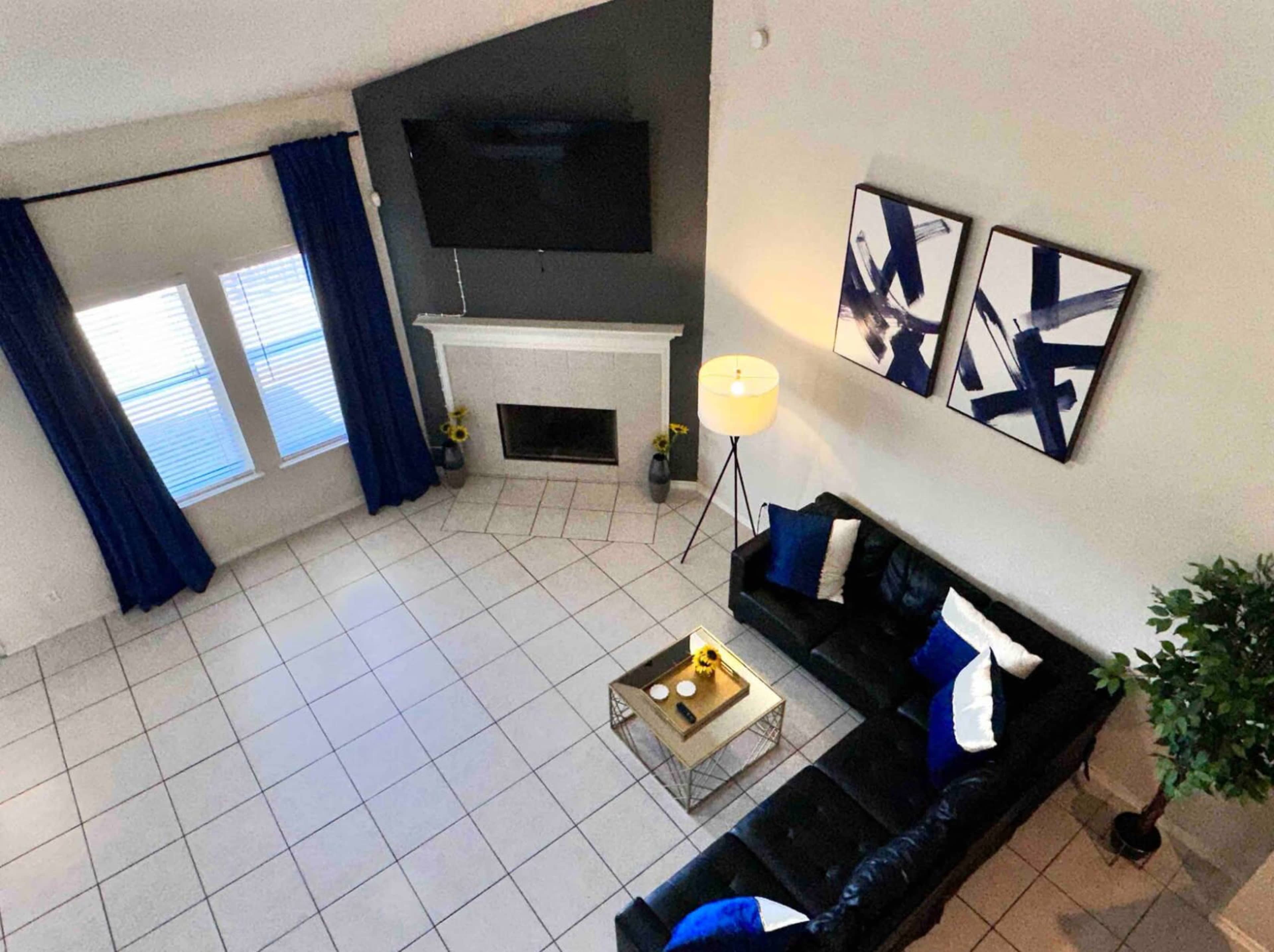 A well-lit living room features a black leather sectional sofa, a glass coffee table, and a fireplace beneath a mounted television, with blue curtains and abstract wall art.