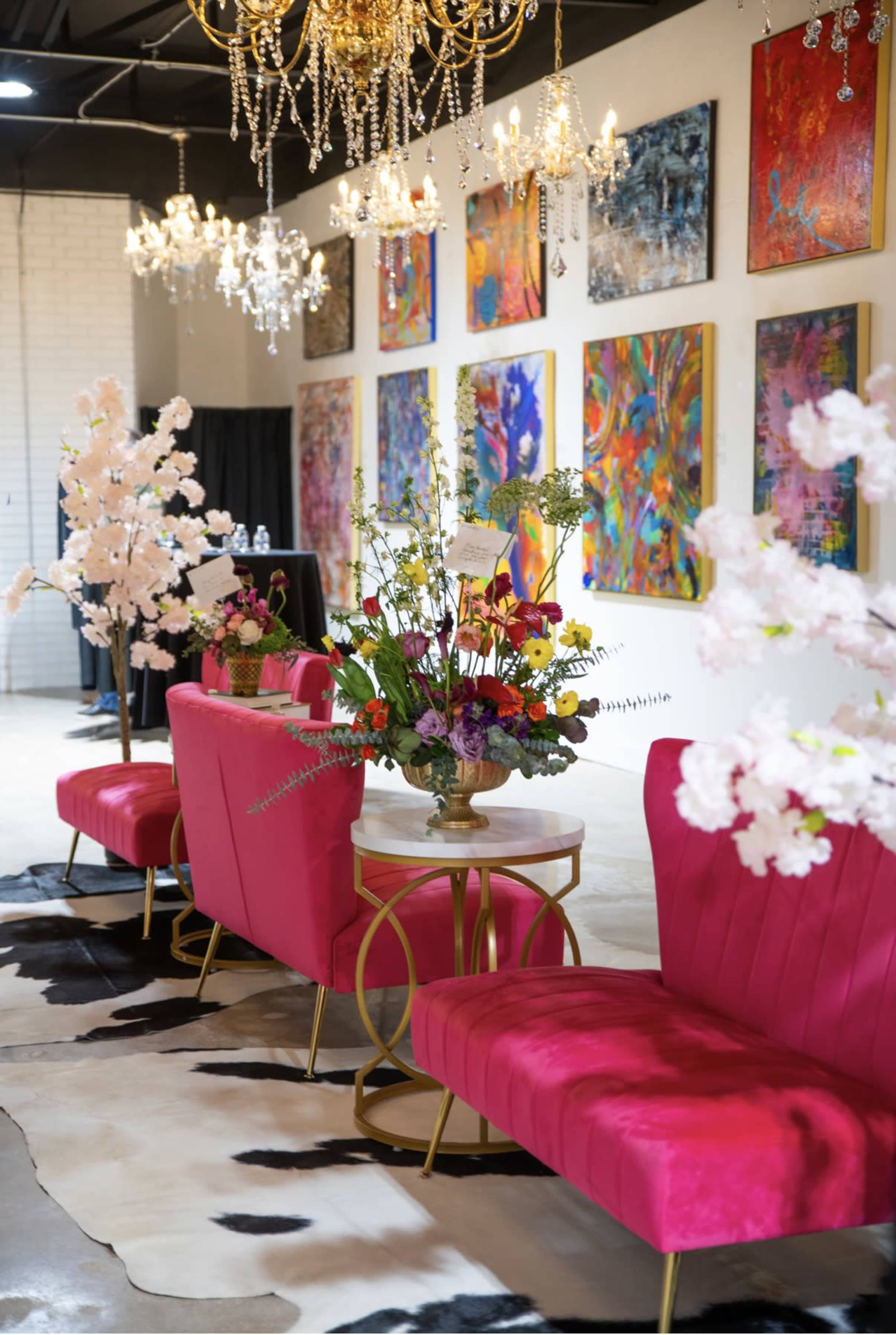 The image shows a modern indoor space featuring pink upholstered seating, decorative floral arrangements, and colorful artwork hanging on the walls, illuminated by elegant chandeliers.