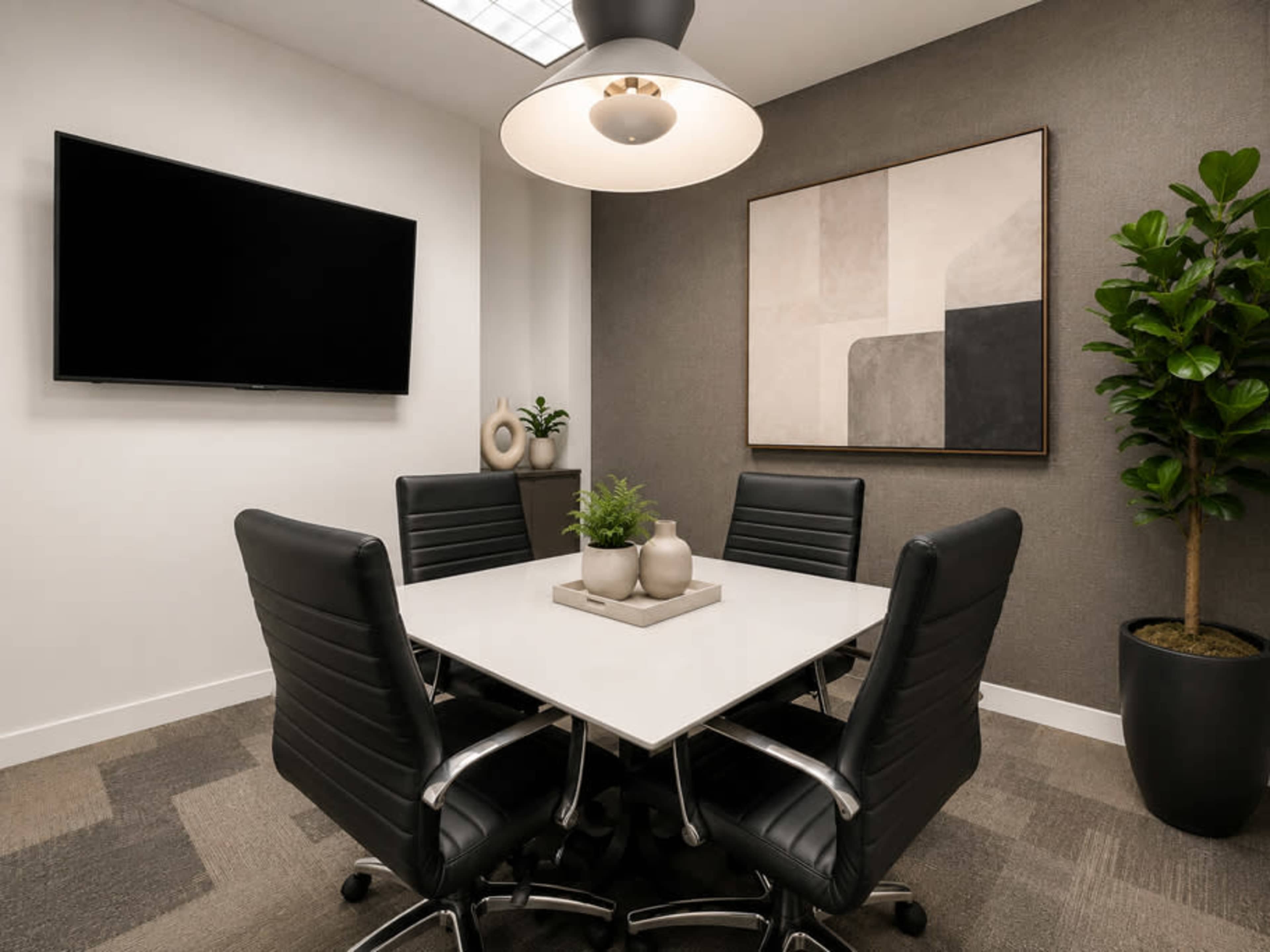 The image shows a modern conference room with a square white table, four black leather chairs, a television mounted on the wall, and a decorative plant in the corner.