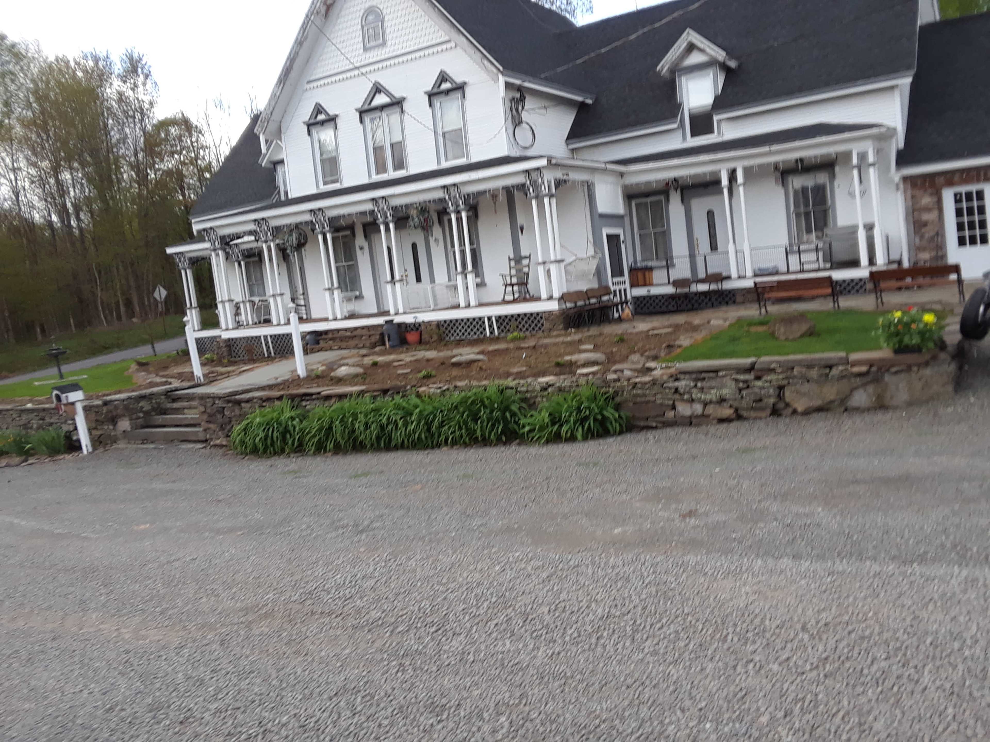 A white, two-story house with a large porch and multiple rocking chairs sits alongside a gravel driveway, surrounded by freshly landscaped gardens.