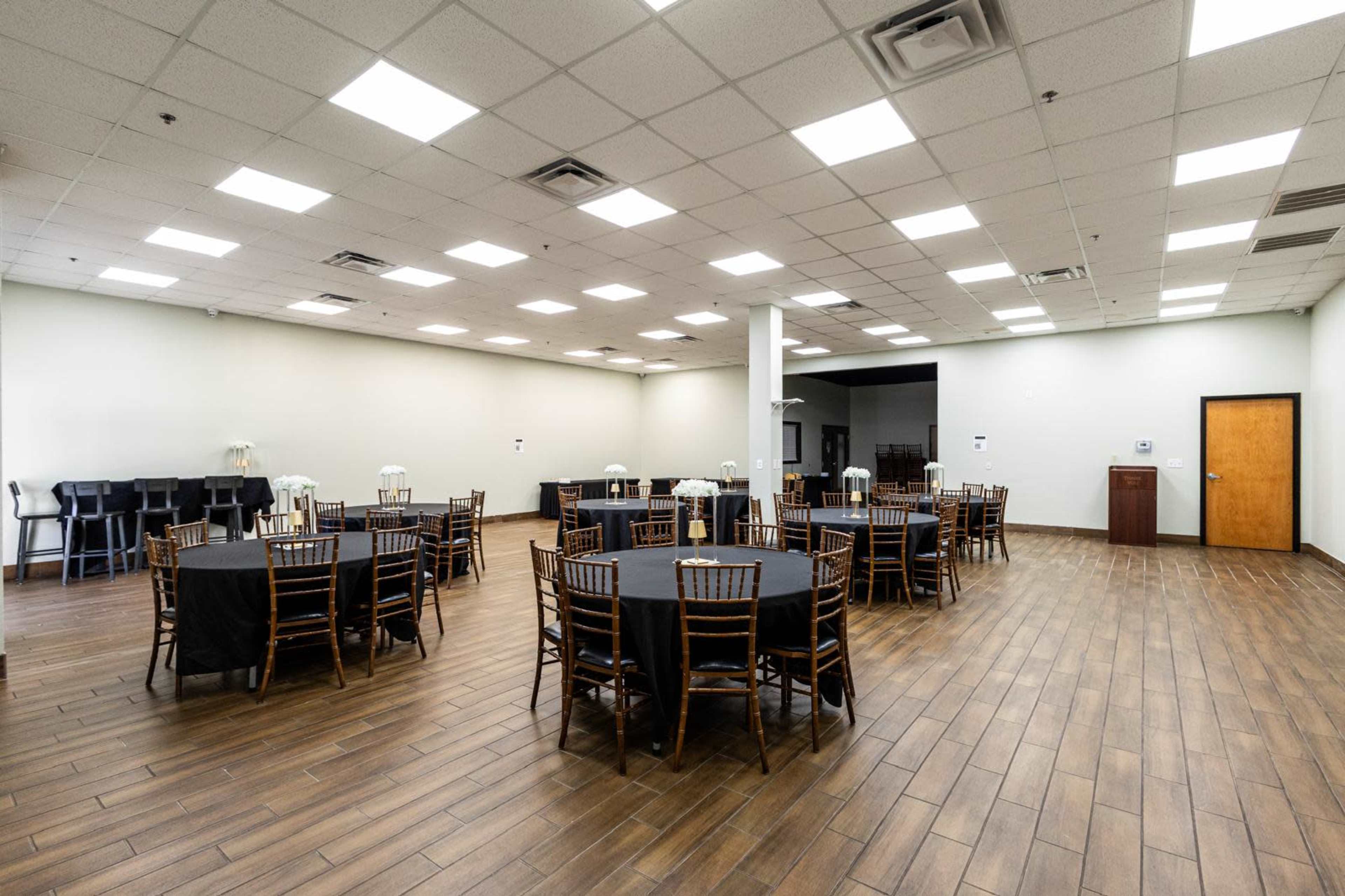 The image shows a spacious event room with several round tables covered in black tablecloths surrounded by gold chiavari chairs, and a wooden floor.