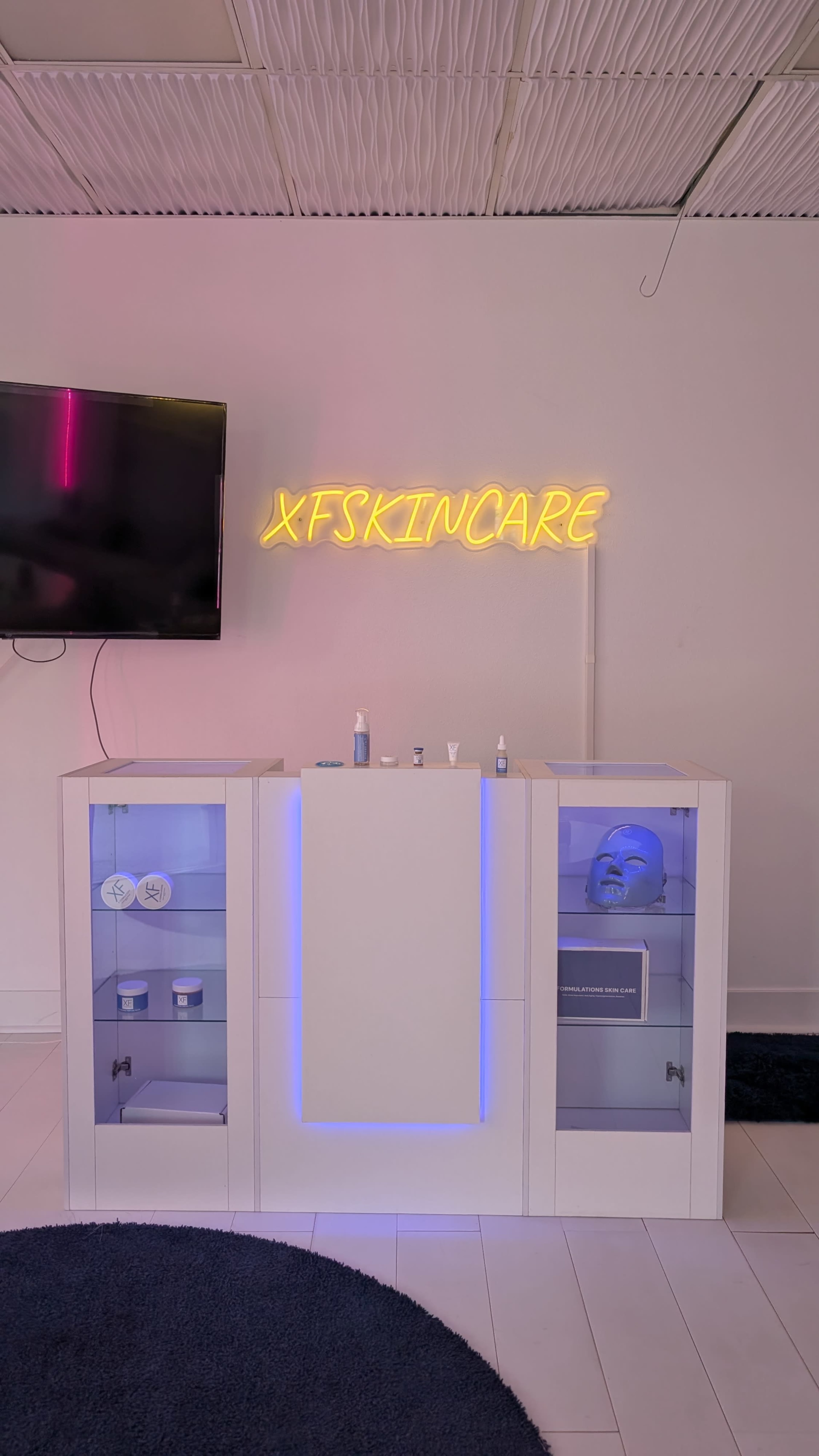 The image shows a modern skincare display featuring a central white cabinet with blue lighting, flanked by glass shelves displaying various skincare products, and a television mounted on the wall above it.
