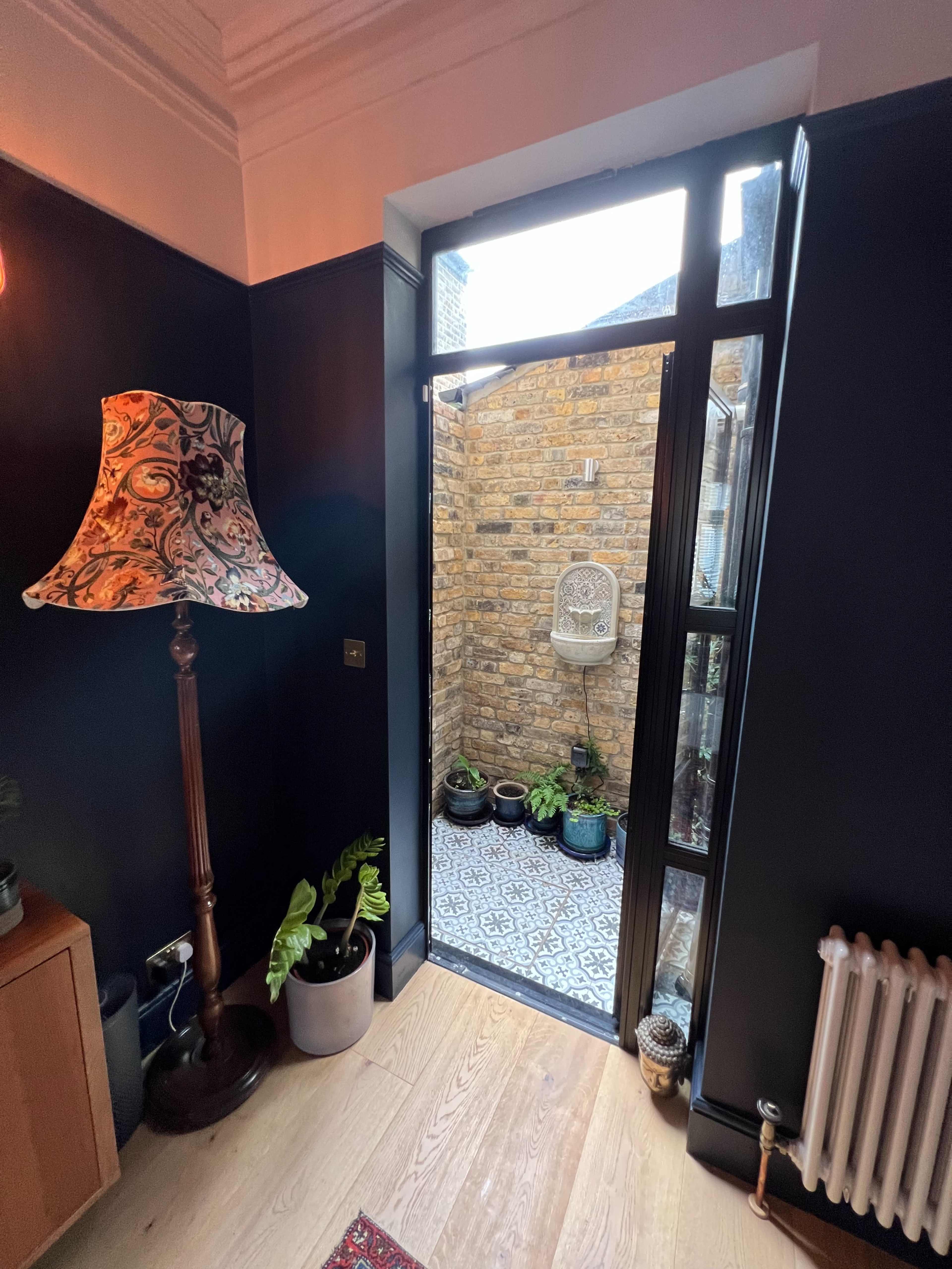 A doorway leading to a small outdoor space with potted plants and a tiled floor, framed by a lamp stand and a wooden cabinet.