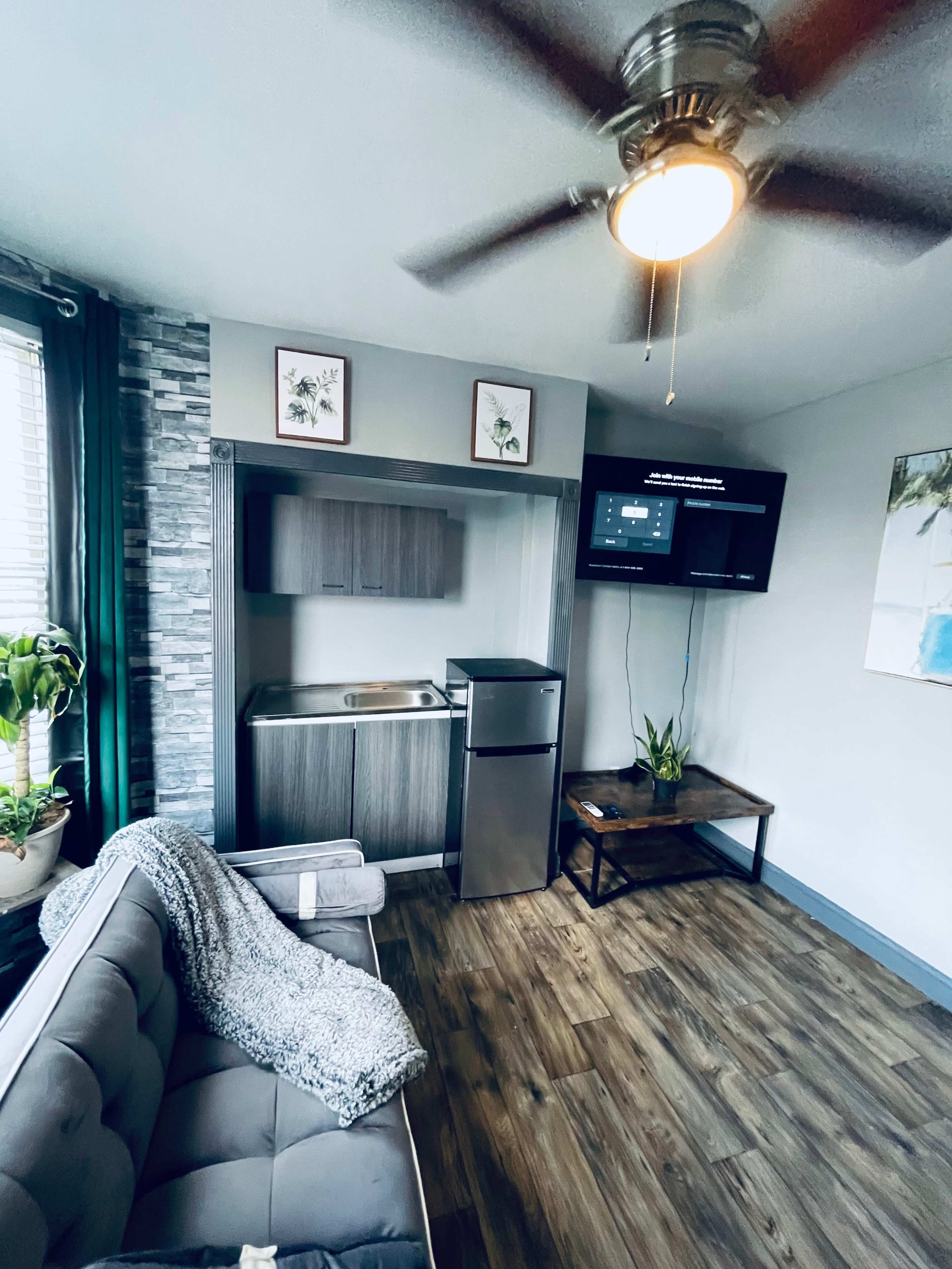 A small, modern living space featuring a couch, a kitchenette with a fridge, and a television mounted on the wall.