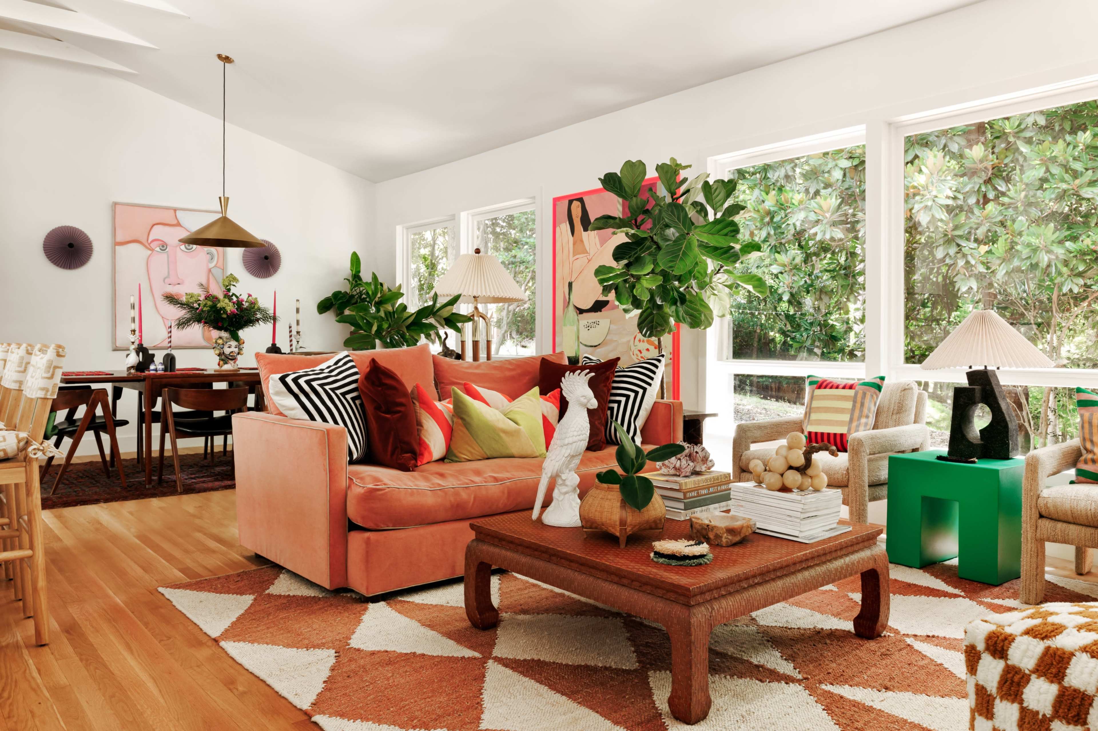 The image shows a living room featuring a peach-colored couch, a coffee table with decorative items, and large windows letting in natural light, complemented by various plants and artwork on the walls.