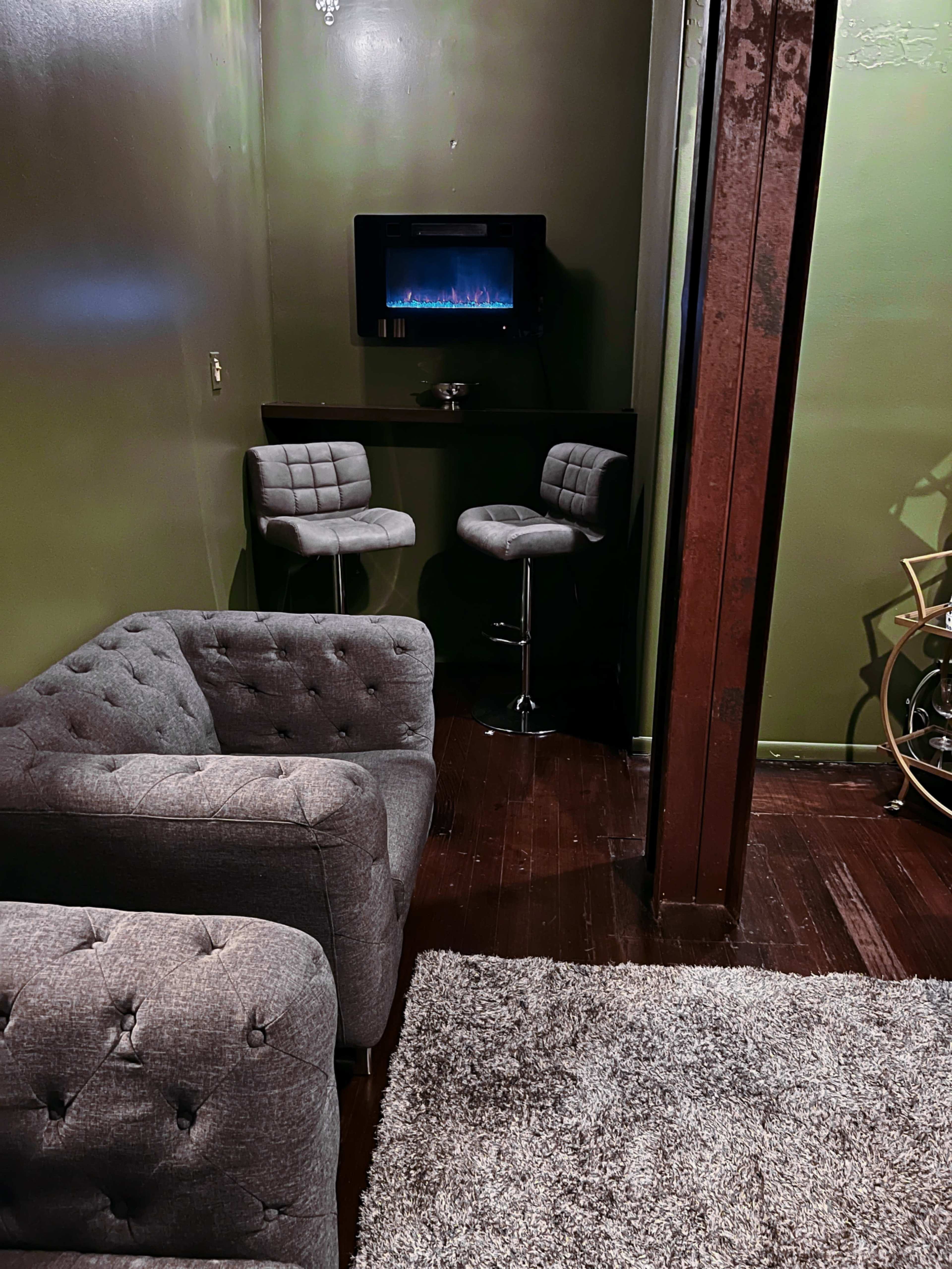 A small lounge area with two barstools and a small TV mounted on the wall, accompanied by a grey tufted sofa and a plush rug on the wooden floor.
