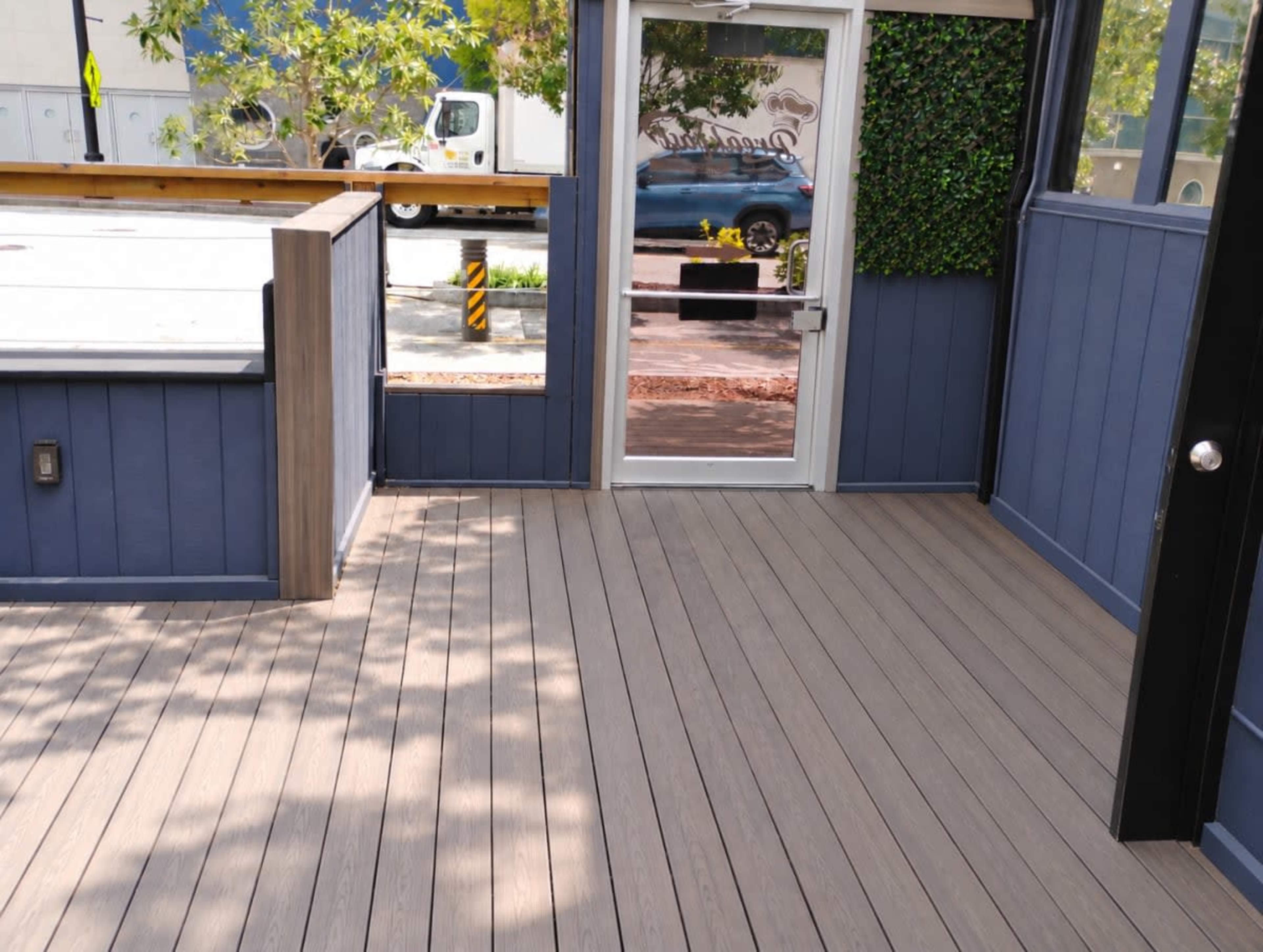 The image shows a wooden deck area leading to a glass door, with greenery visible near the entrance.