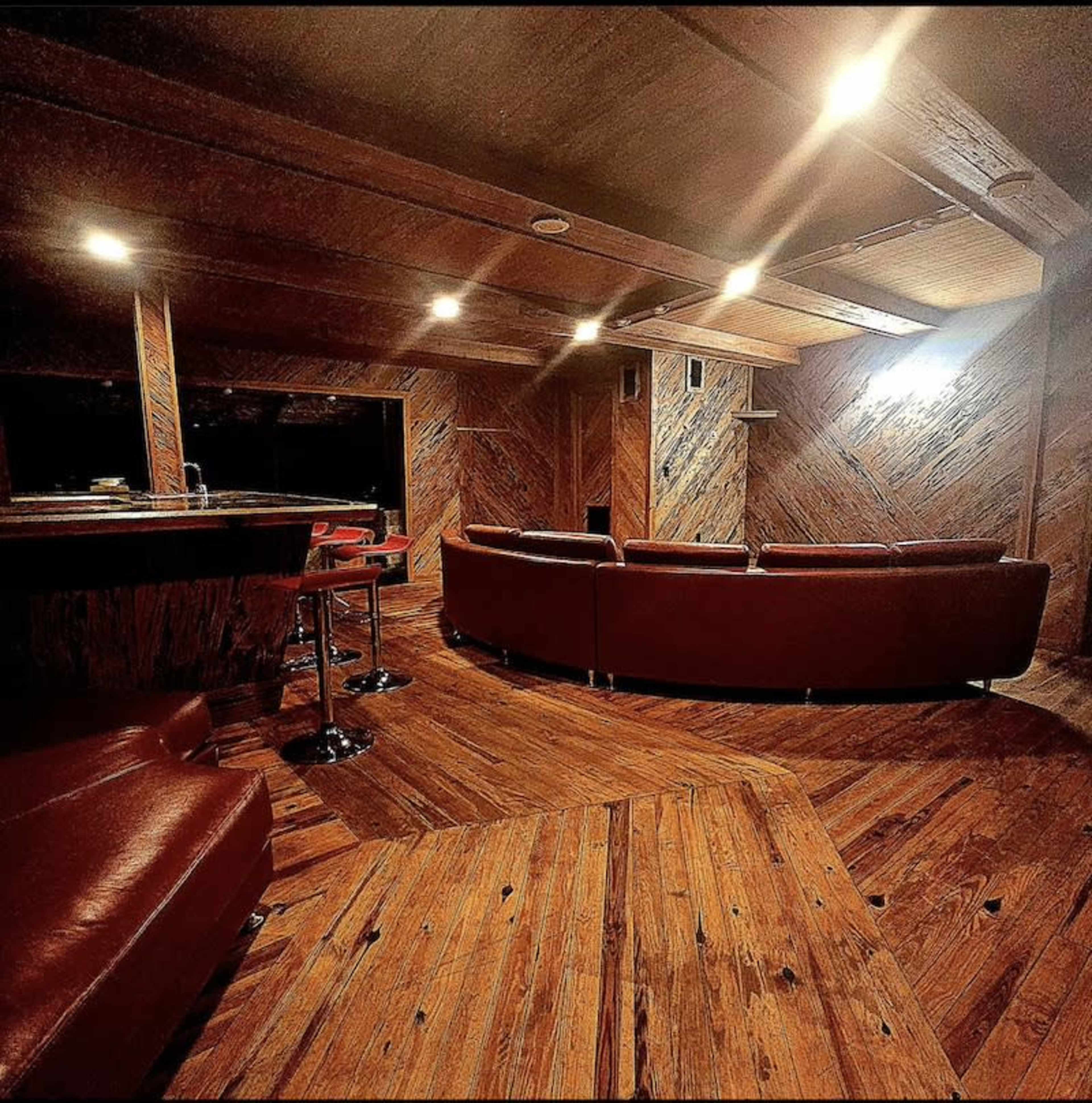 The image shows a wooden-paneled room with a circular seating arrangement, a bar area, and dim overhead lighting.