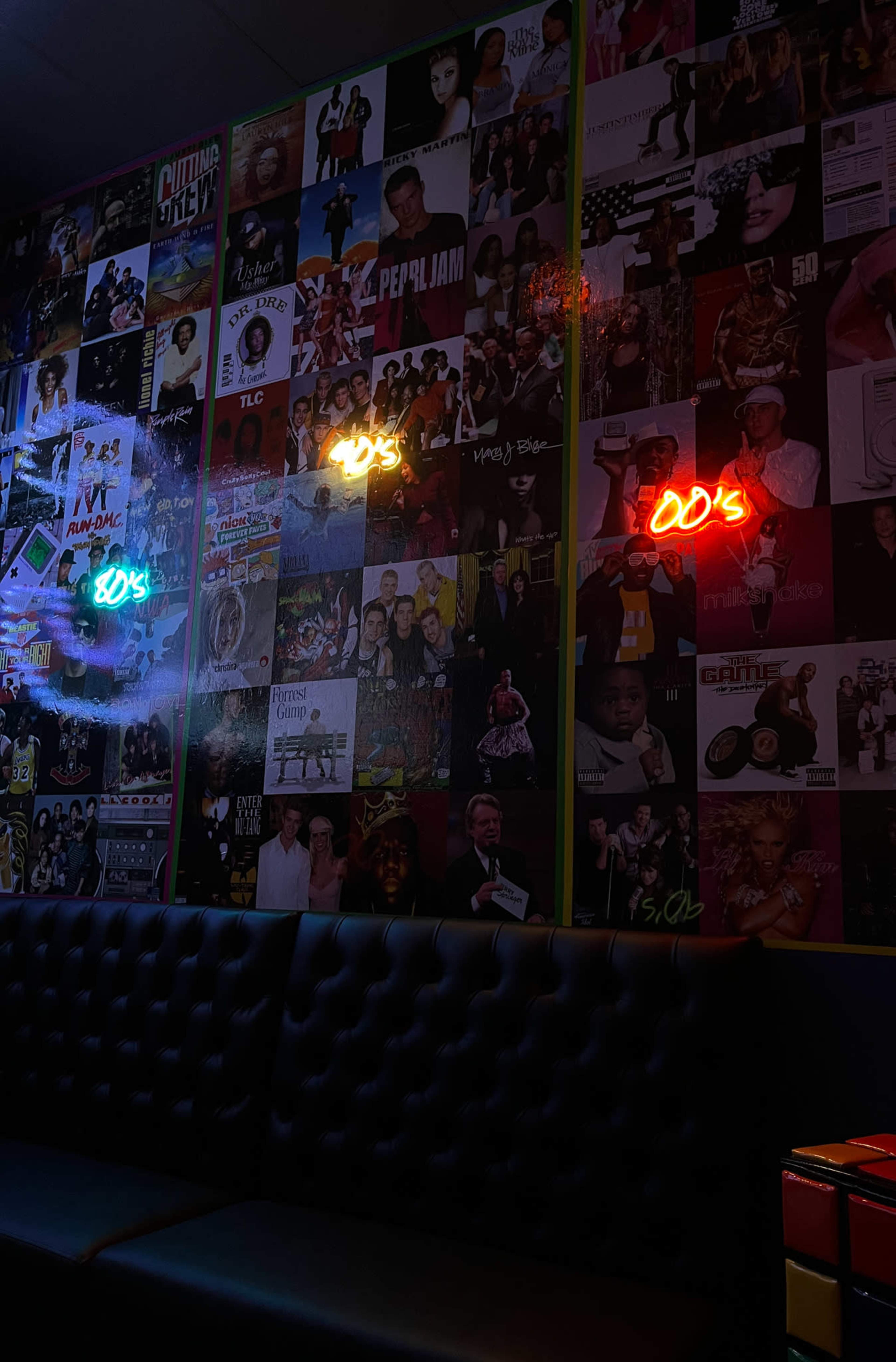 A dark room with a black couch and walls covered in colorful posters from various artists, along with neon signs that read "90s" and "00s."