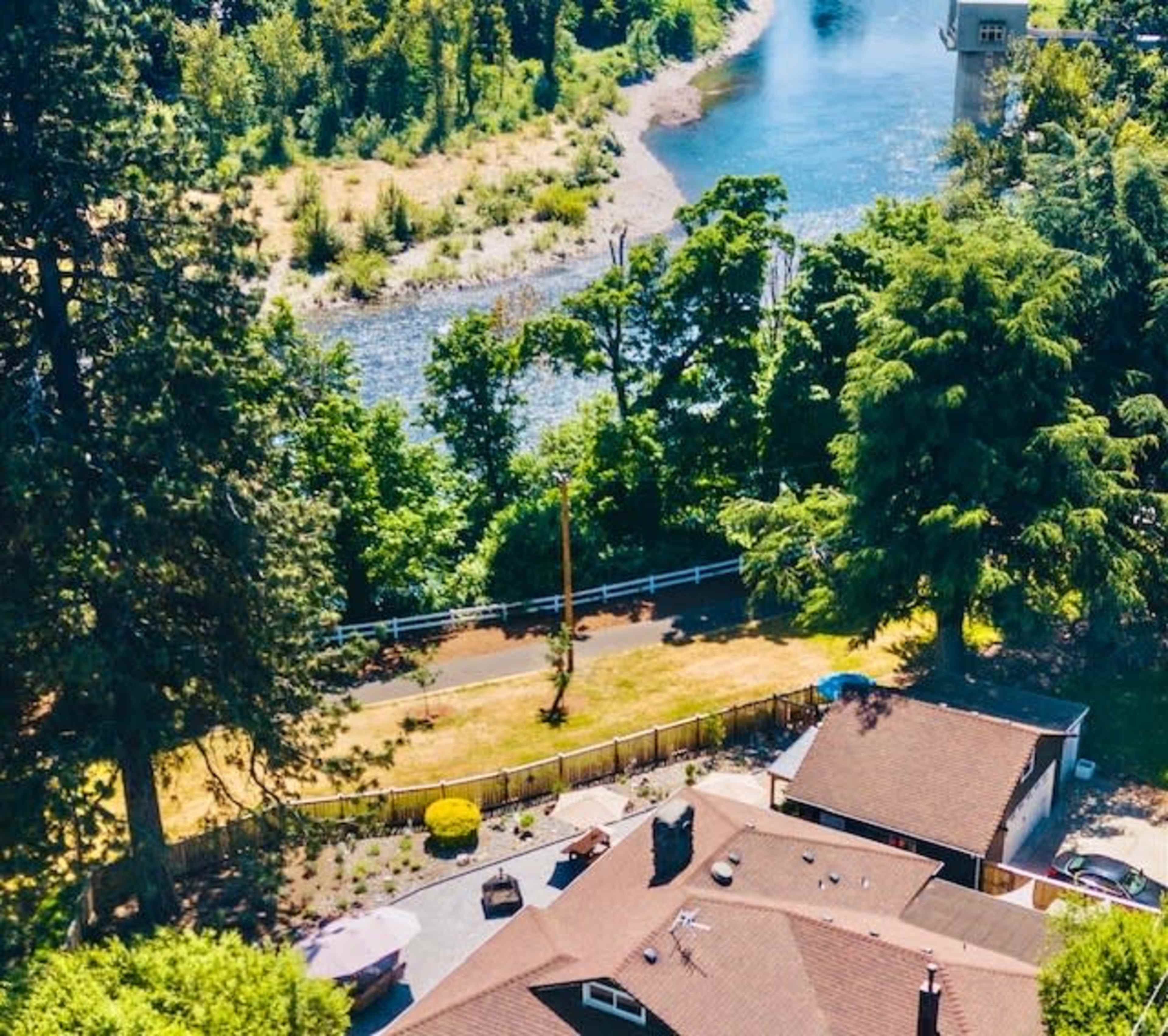 The image shows a river bordered by trees and a residential area with a brown roof and a nearby path.
