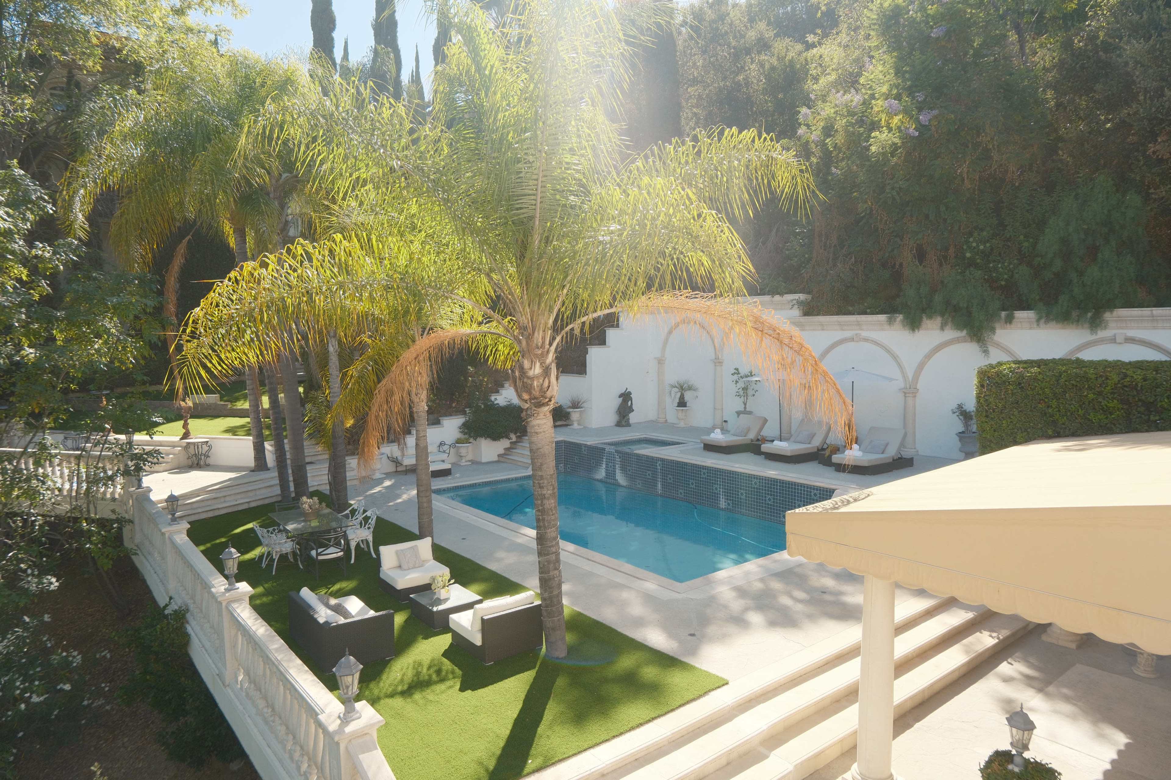 The image shows a luxury backyard featuring a swimming pool surrounded by palm trees, lounge chairs, and an outdoor seating area.