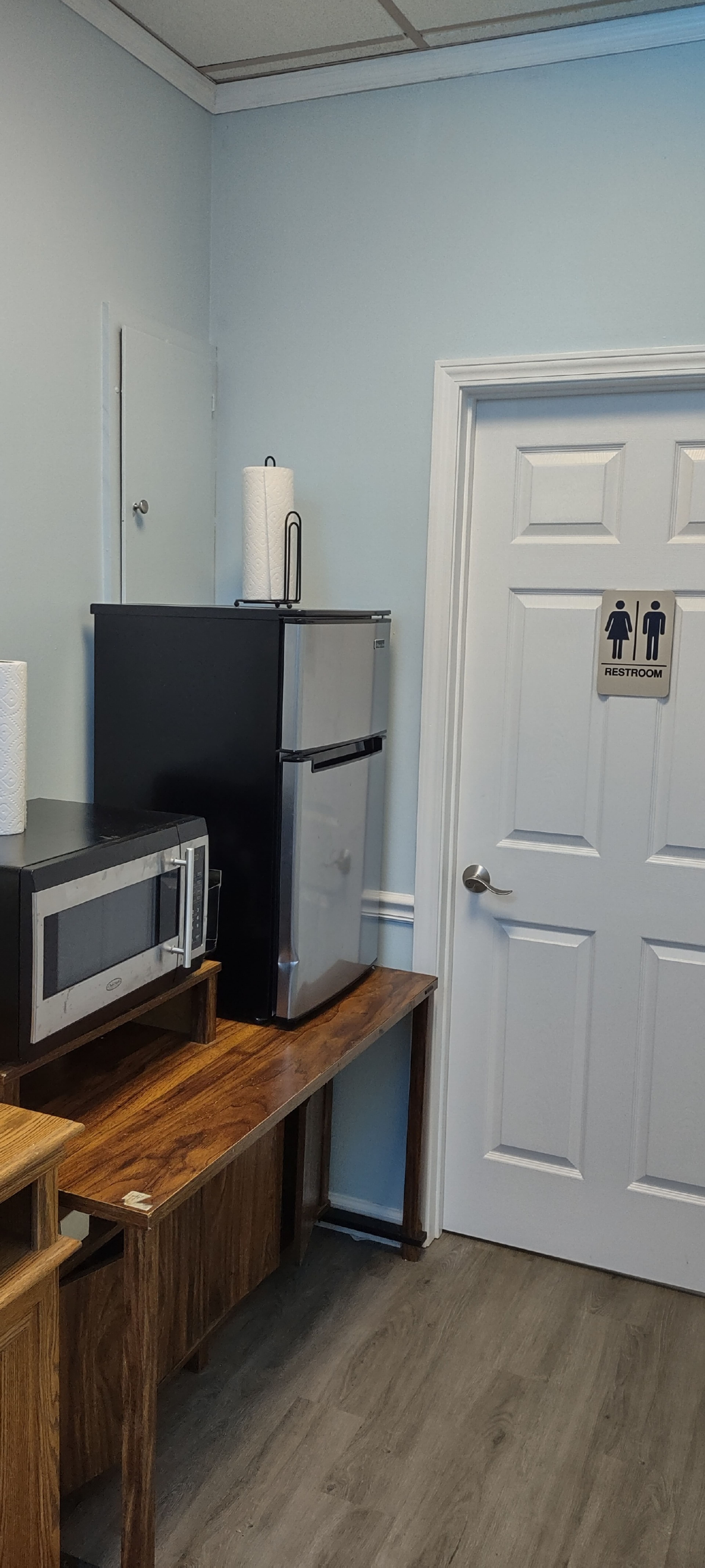 The image shows a small break room corner with a black refrigerator, a microwave on a wooden table, and a door labeled "RESTROOM" beside it.
