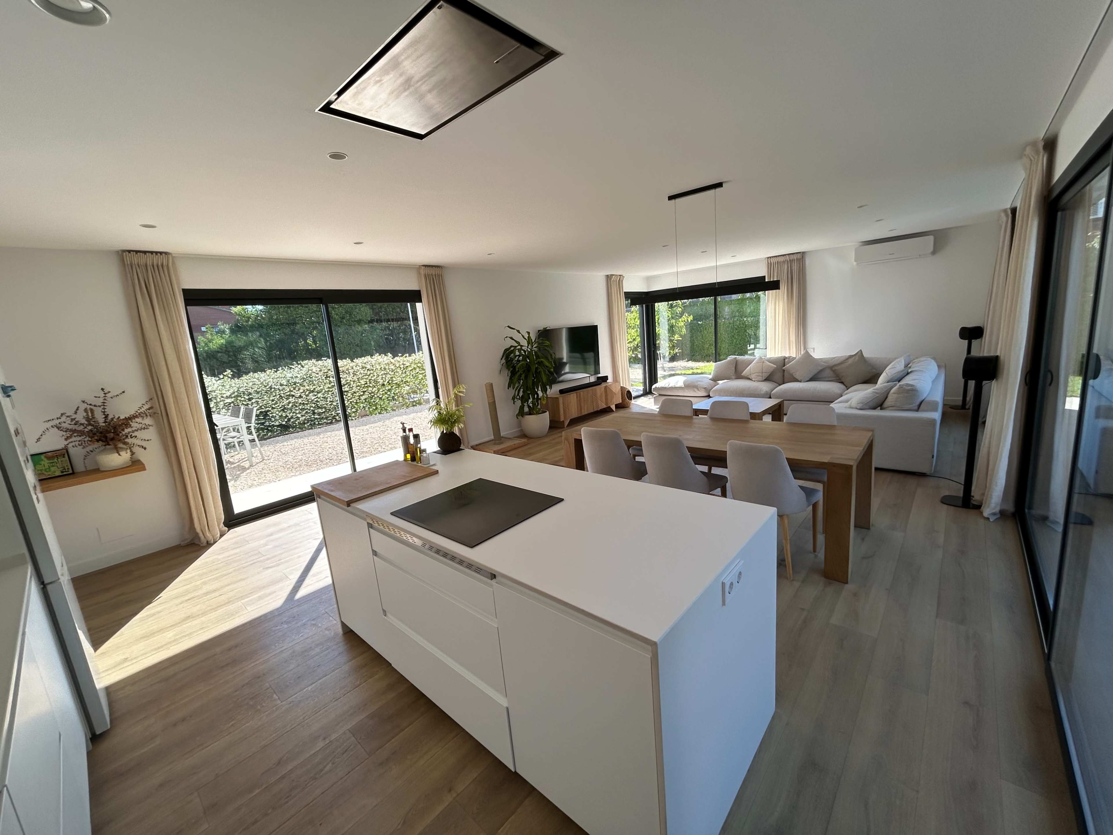 The image shows a modern open-concept living space featuring a kitchen island, dining table, and a lounge area with large windows overlooking a garden.
