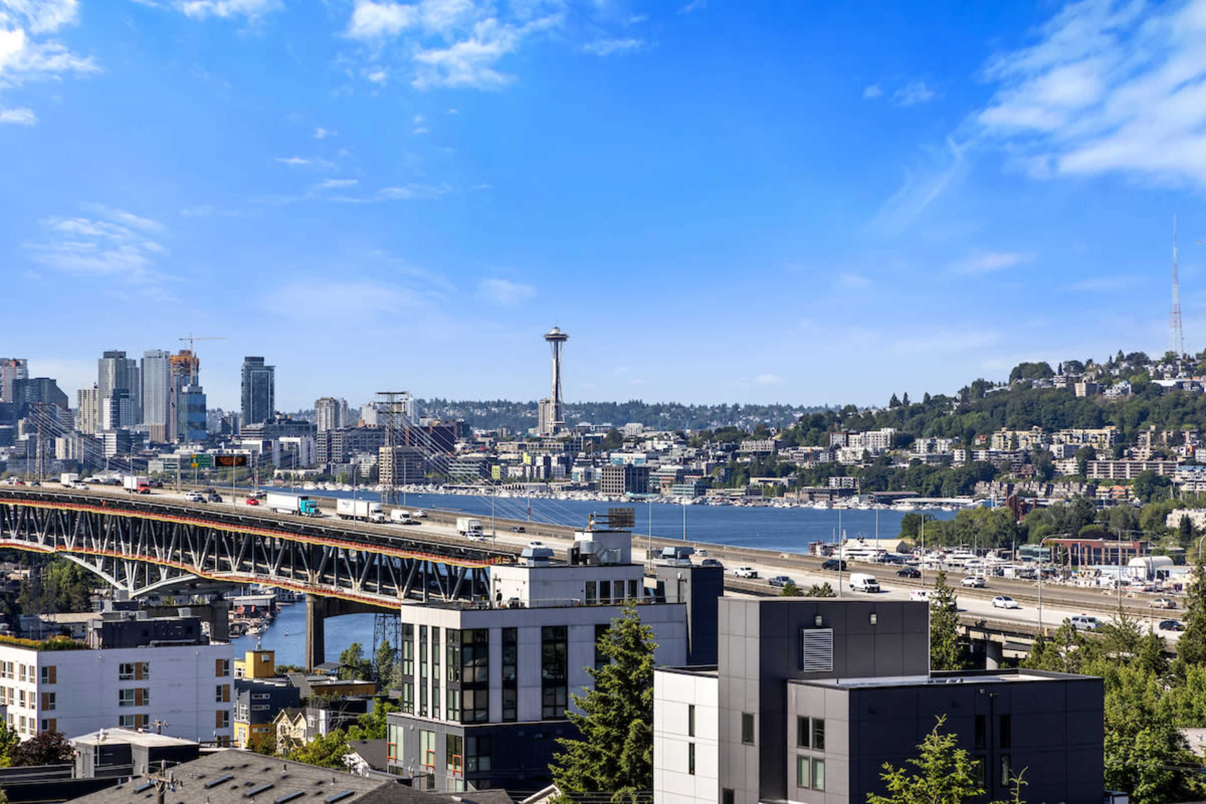 The image shows a view of Seattle, featuring the Space Needle, a bridge, and the skyline with buildings under a clear blue sky.