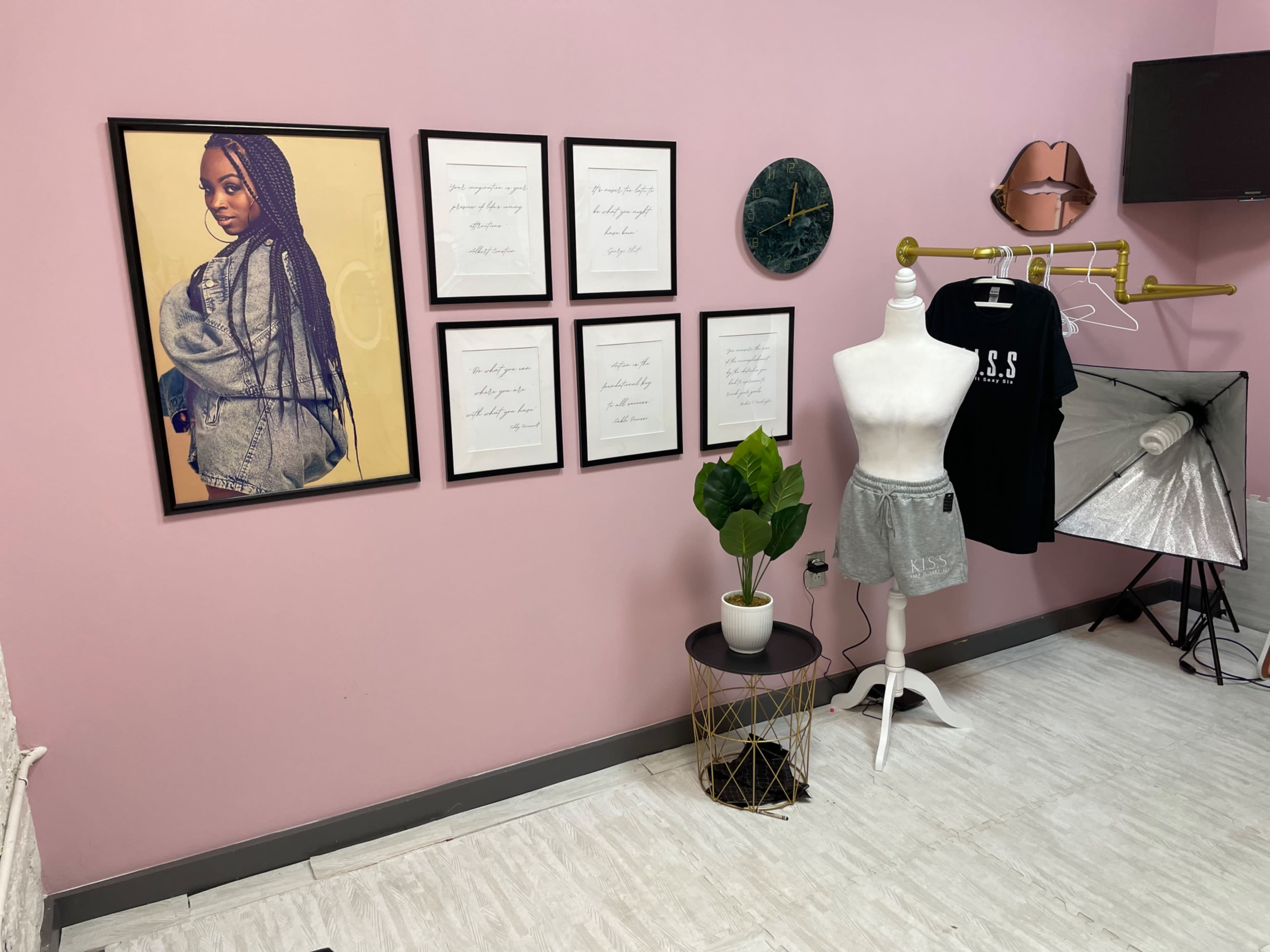 The image shows a room with pink walls, featuring a framed portrait on the left, a series of framed texts, a mannequin displaying a black shirt, a potted plant, and a photography lighting setup.