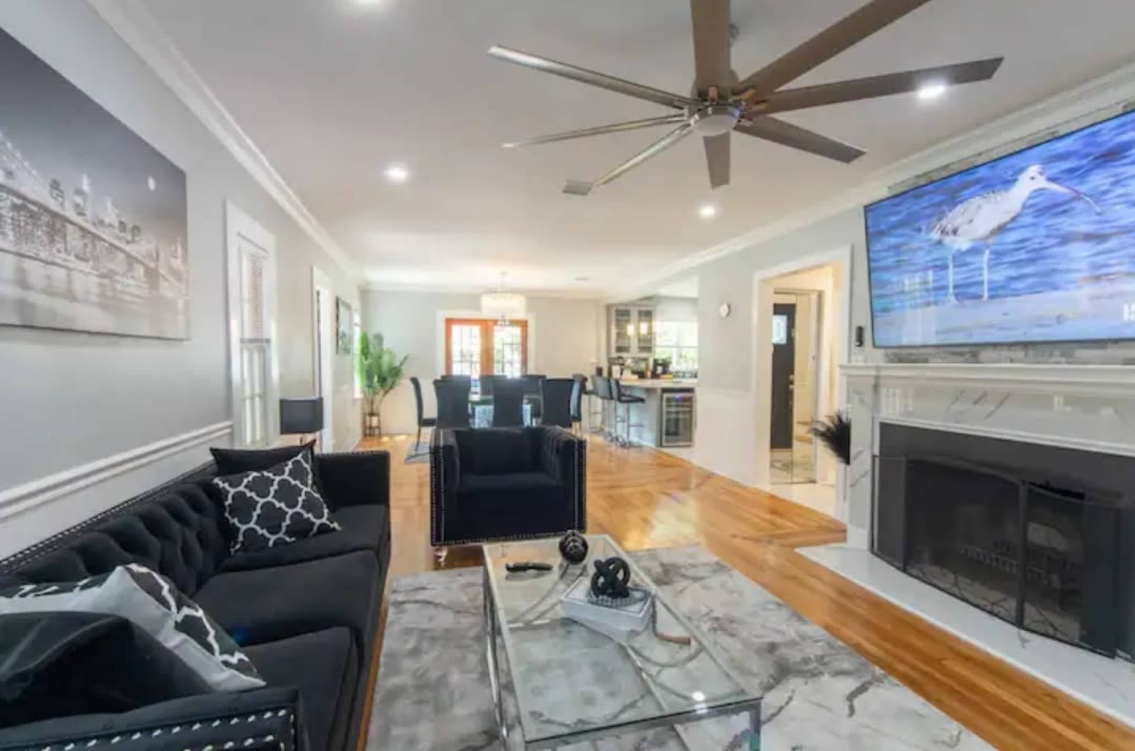 The living room features a large black sofa, a glass coffee table, and a mounted TV above a fireplace, with a view into a dining area and modern kitchen.