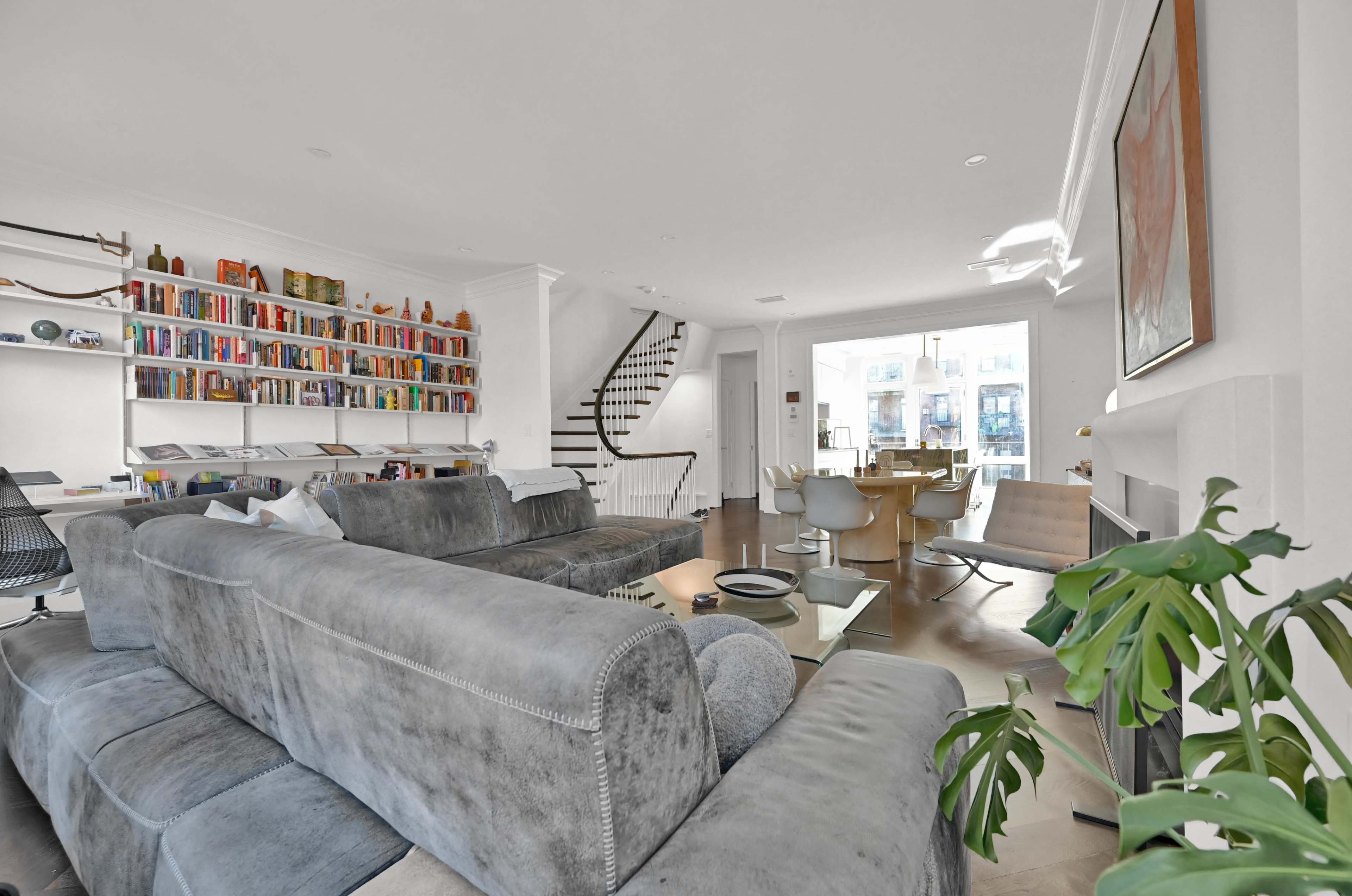 A spacious living area features a large sectional sofa, a bookshelf filled with numerous books, and a dining table next to a window leading to a balcony.