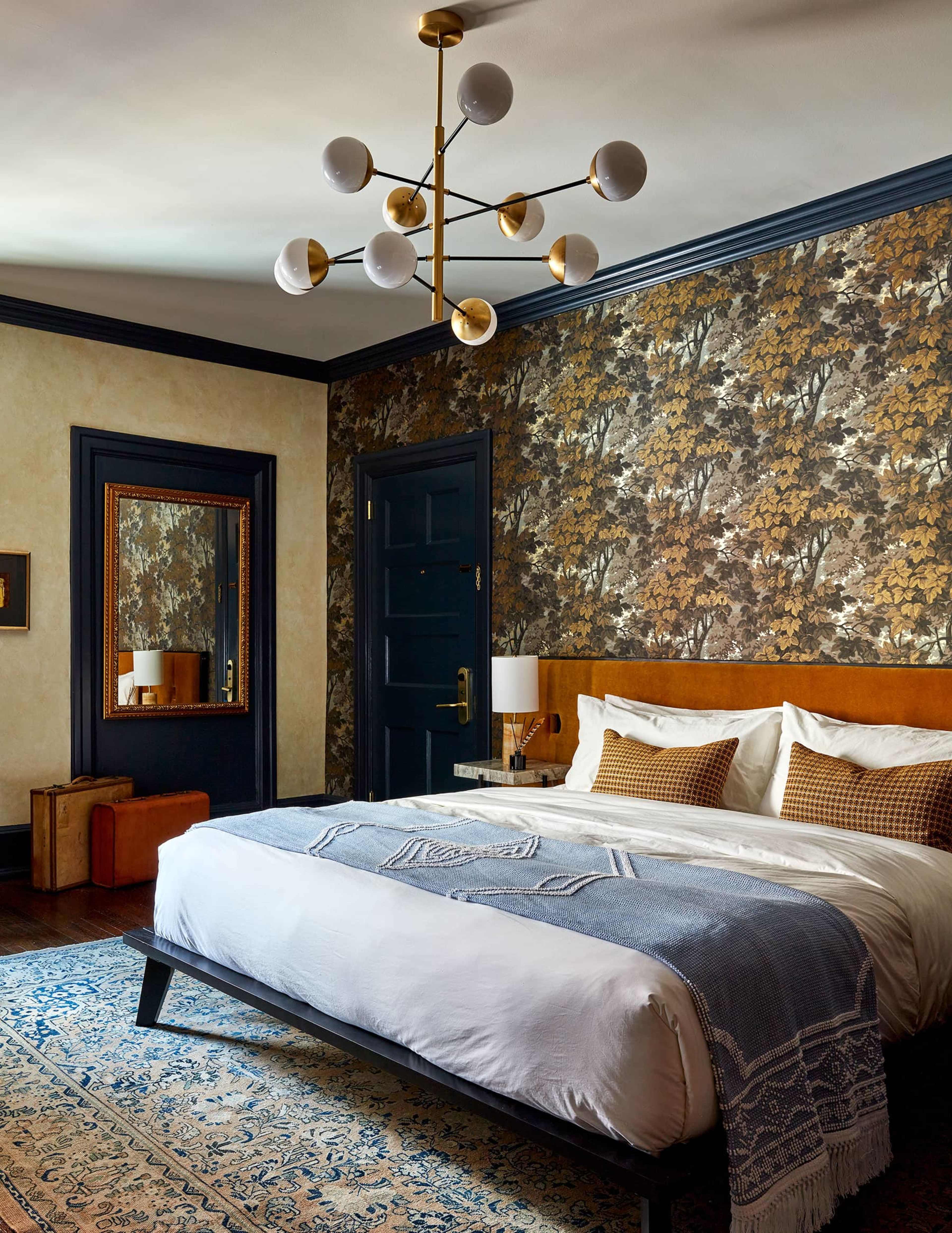 The bedroom features a large bed with white linens and a blue throw blanket, set against a wallpapered wall and illuminated by a modern chandelier.