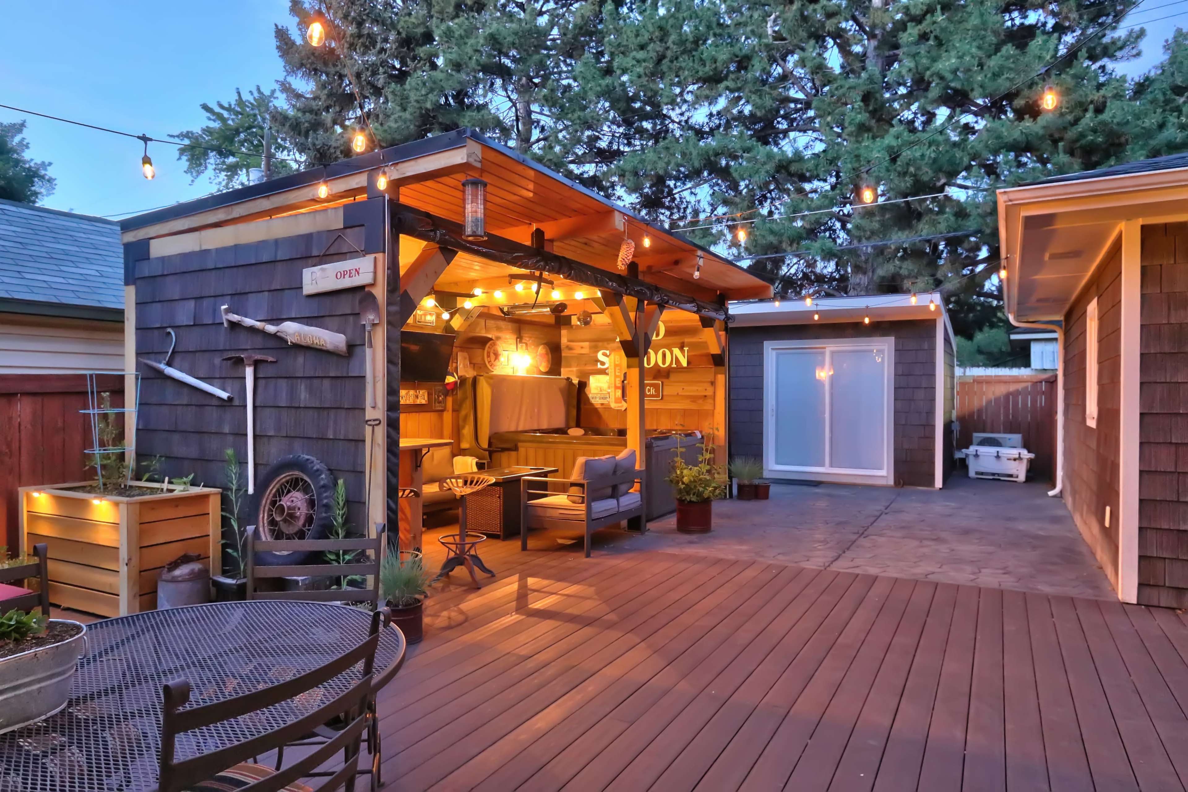 A wooden deck features a well-lit outdoor lounge area with a covered seating space and decorative string lights, alongside a small dining table.