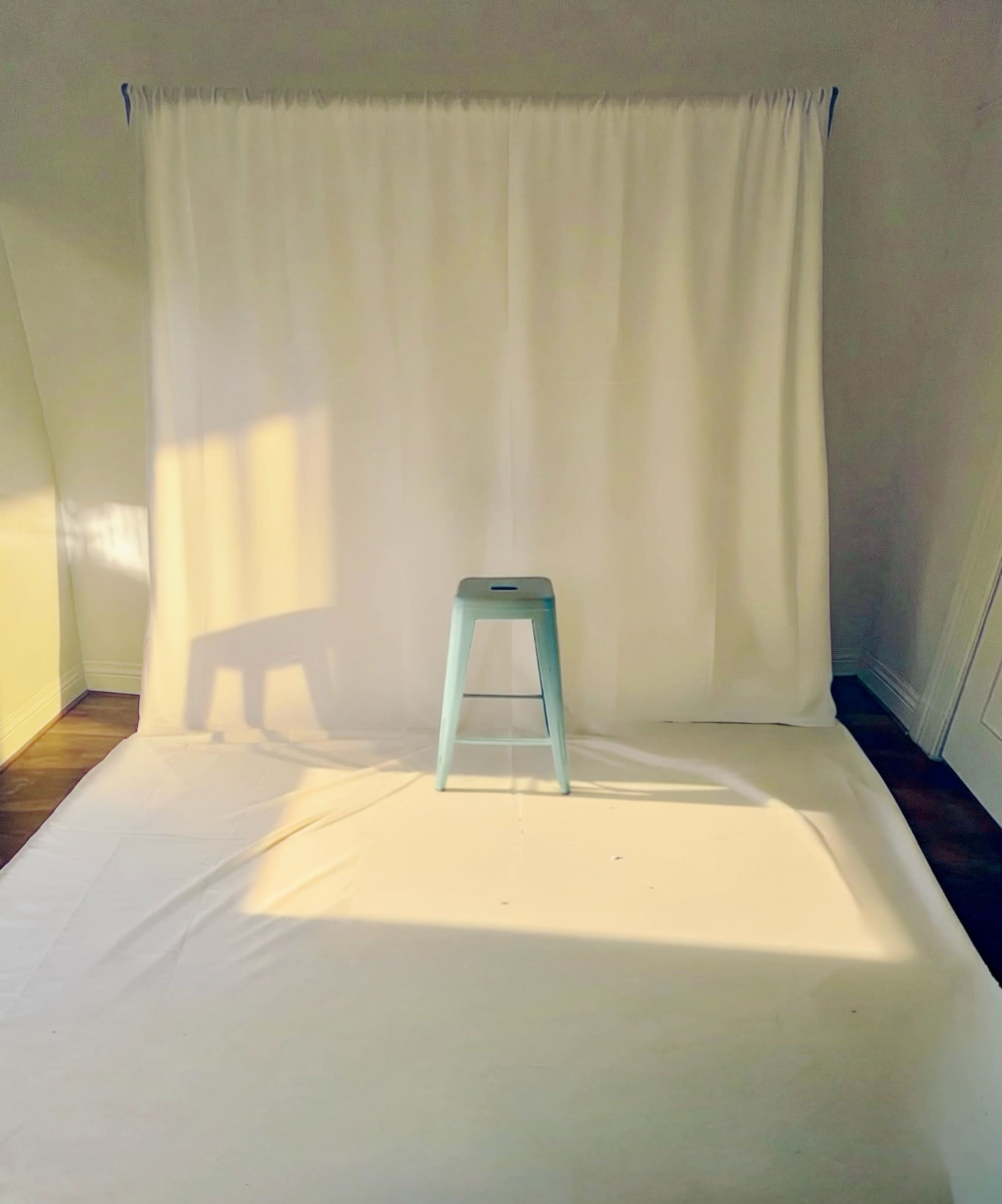 The image shows a simple studio setup with a light blue stool centered in front of a white backdrop and soft natural light casting shadows on the ground.
