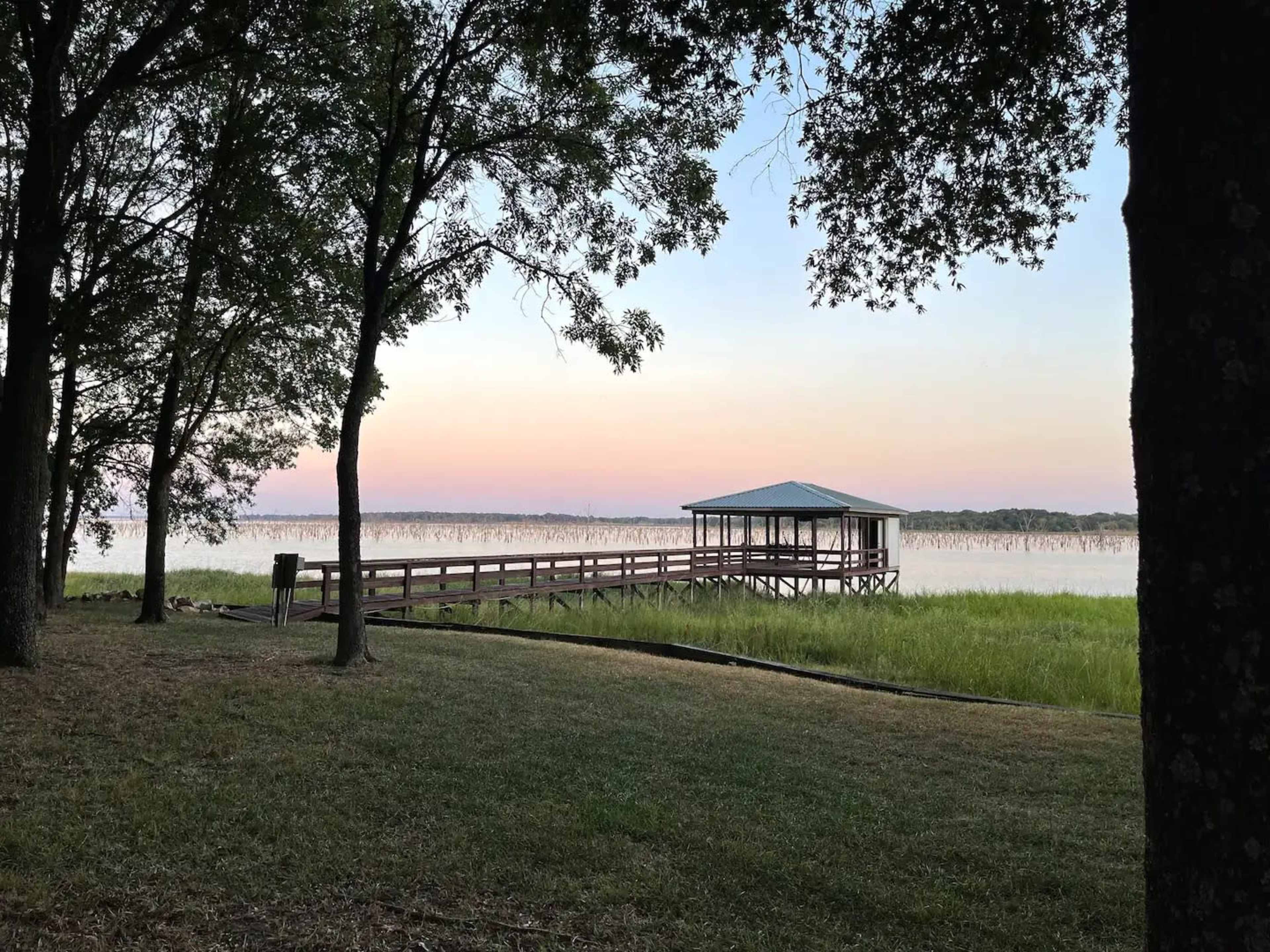 A wooden dock extends over a calm body of water surrounded by trees at dusk.
