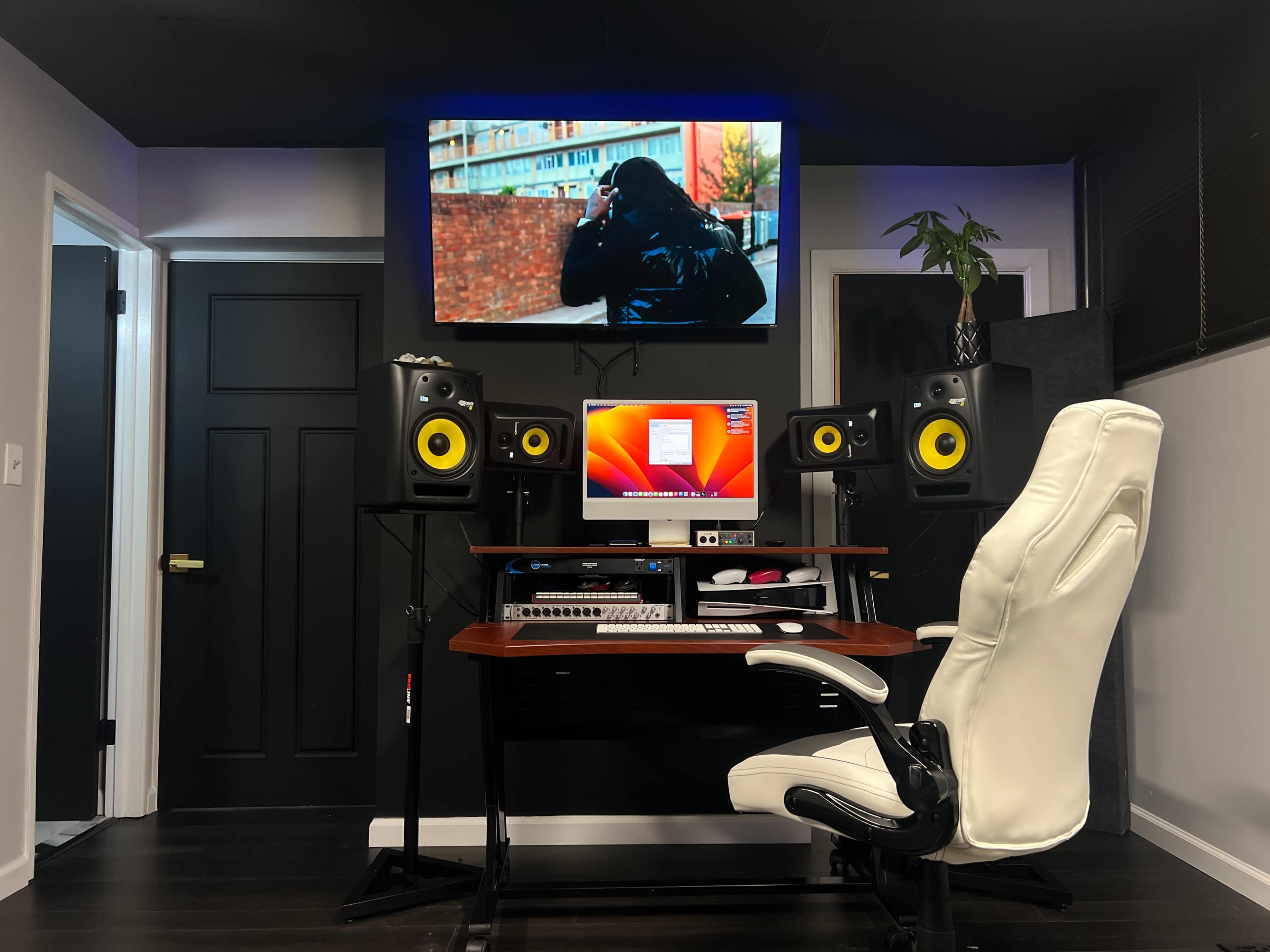 A modern music production studio with a computer setup, studio monitors, and a large screen displaying a person outside.