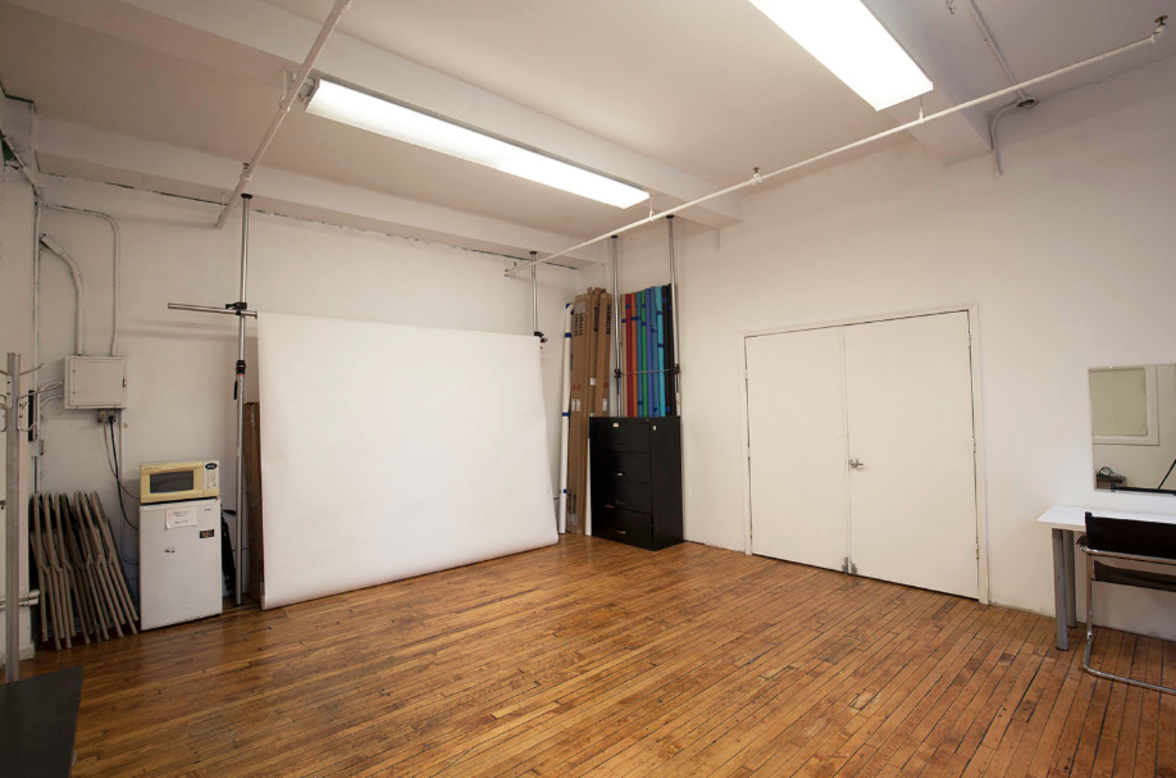 An empty studio space with a white backdrop, wooden floors, and various equipment along the walls.