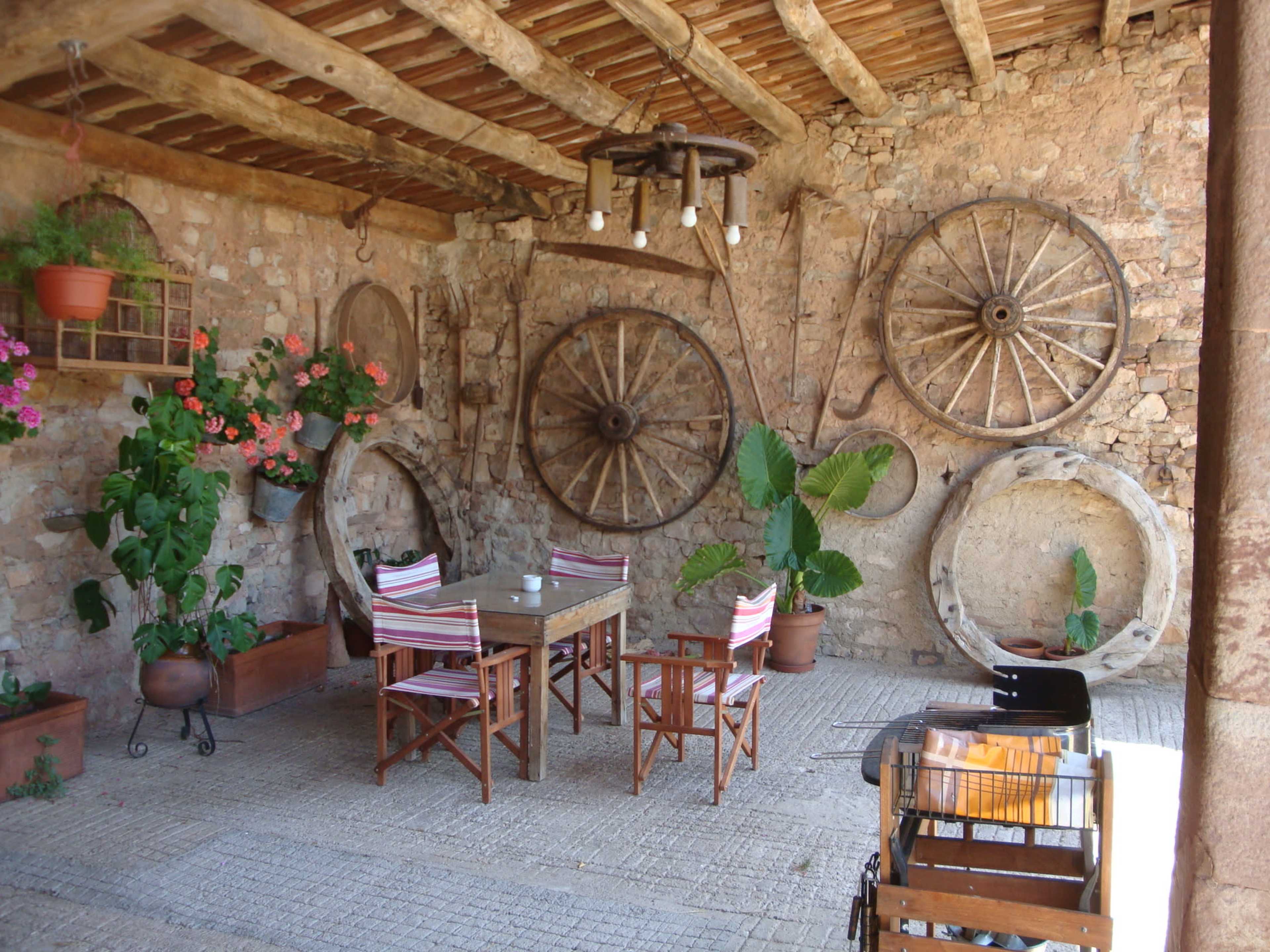 The image shows a rustic dining area with wooden furniture, large decorative wheels on the stone wall, and potted plants.