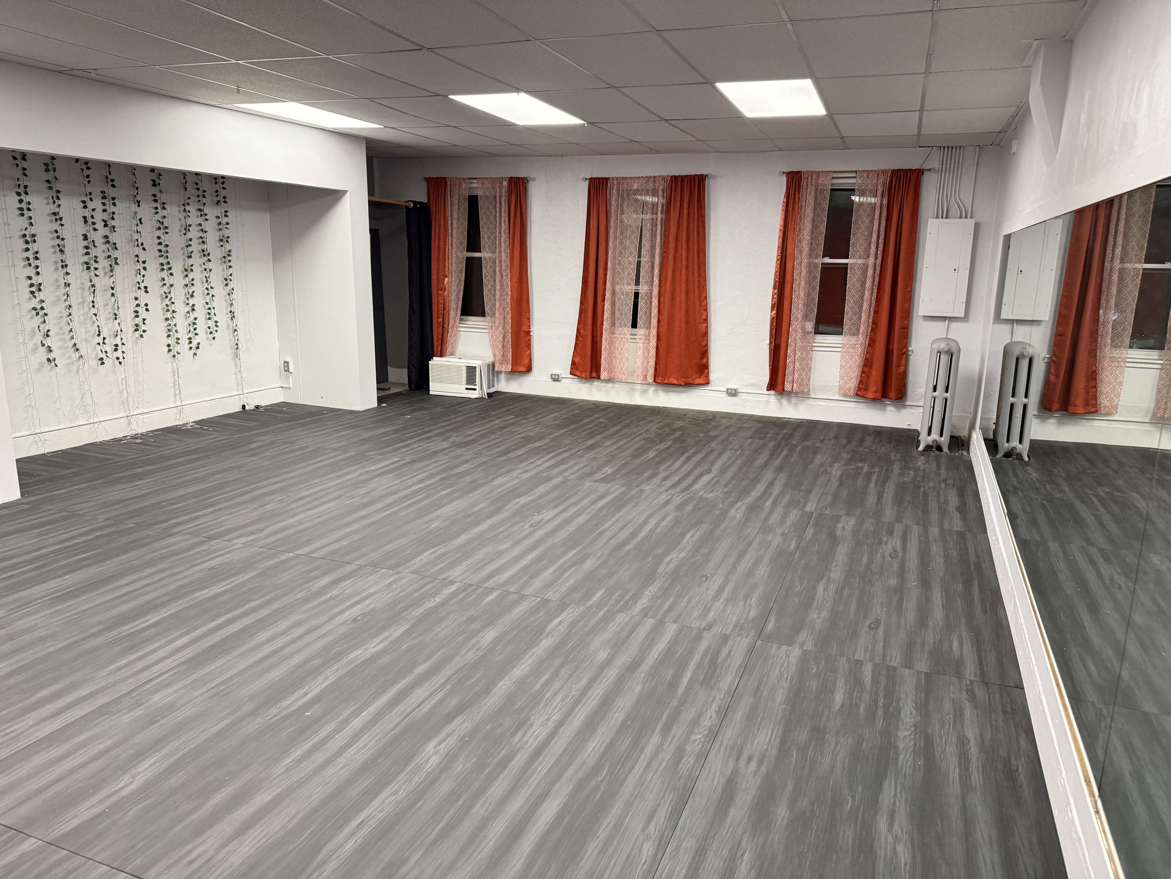 The image shows an empty room with gray flooring, white walls, and large windows covered with orange curtains.