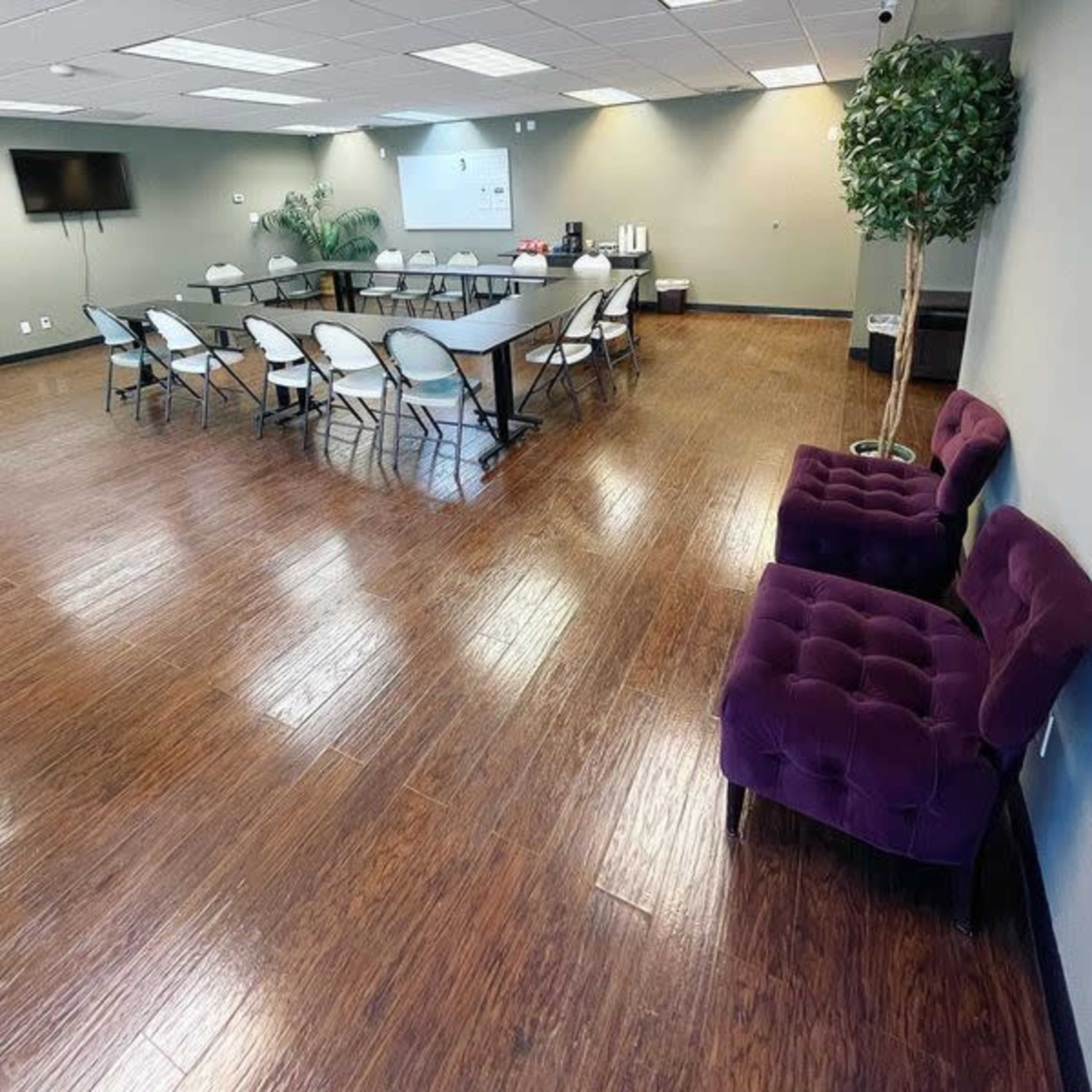 A spacious meeting room features a long table with surrounding folding chairs and two purple armchairs in one corner.