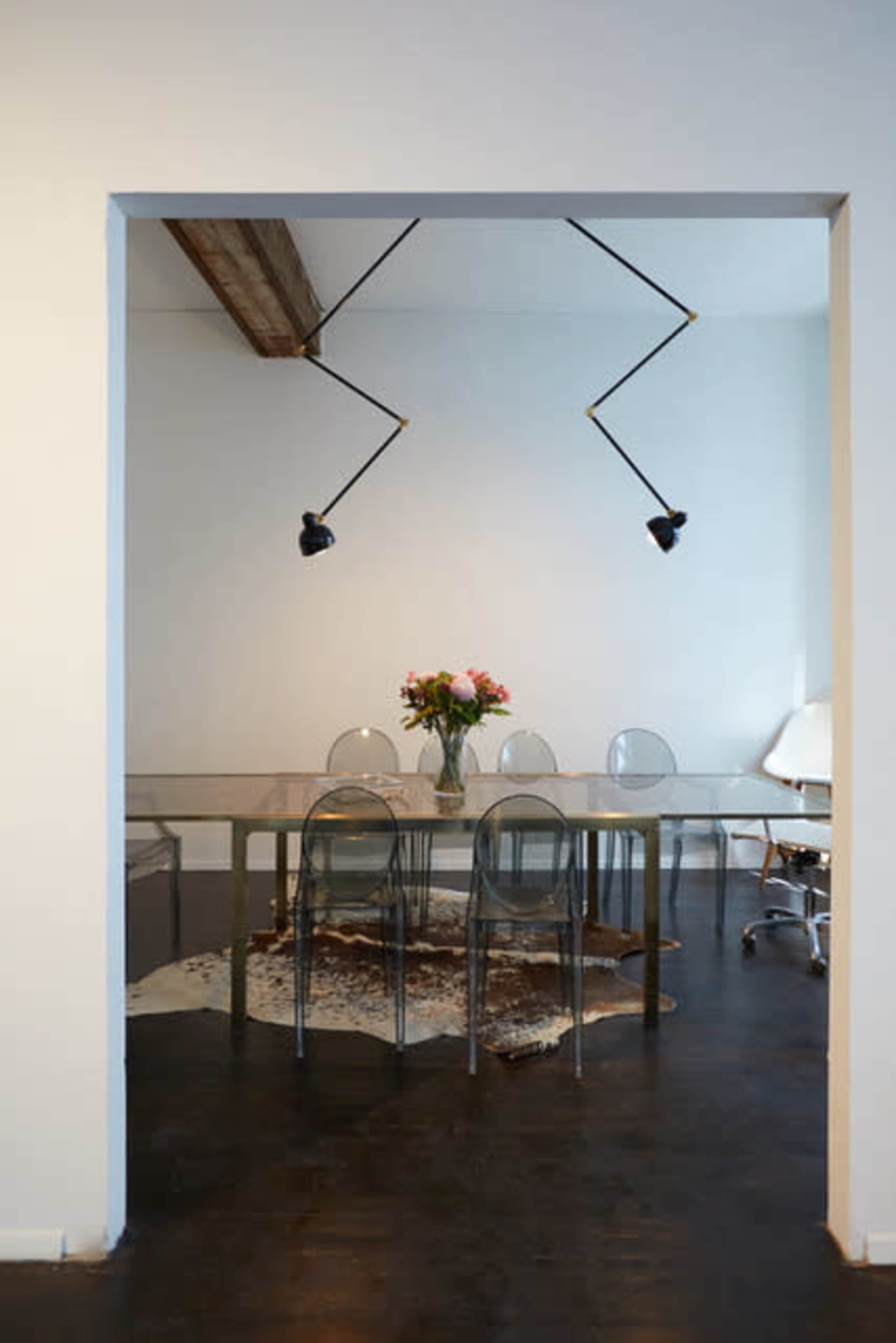 A glass dining table with transparent chairs is set in a room featuring a vase of flowers and stylish lighting fixtures.