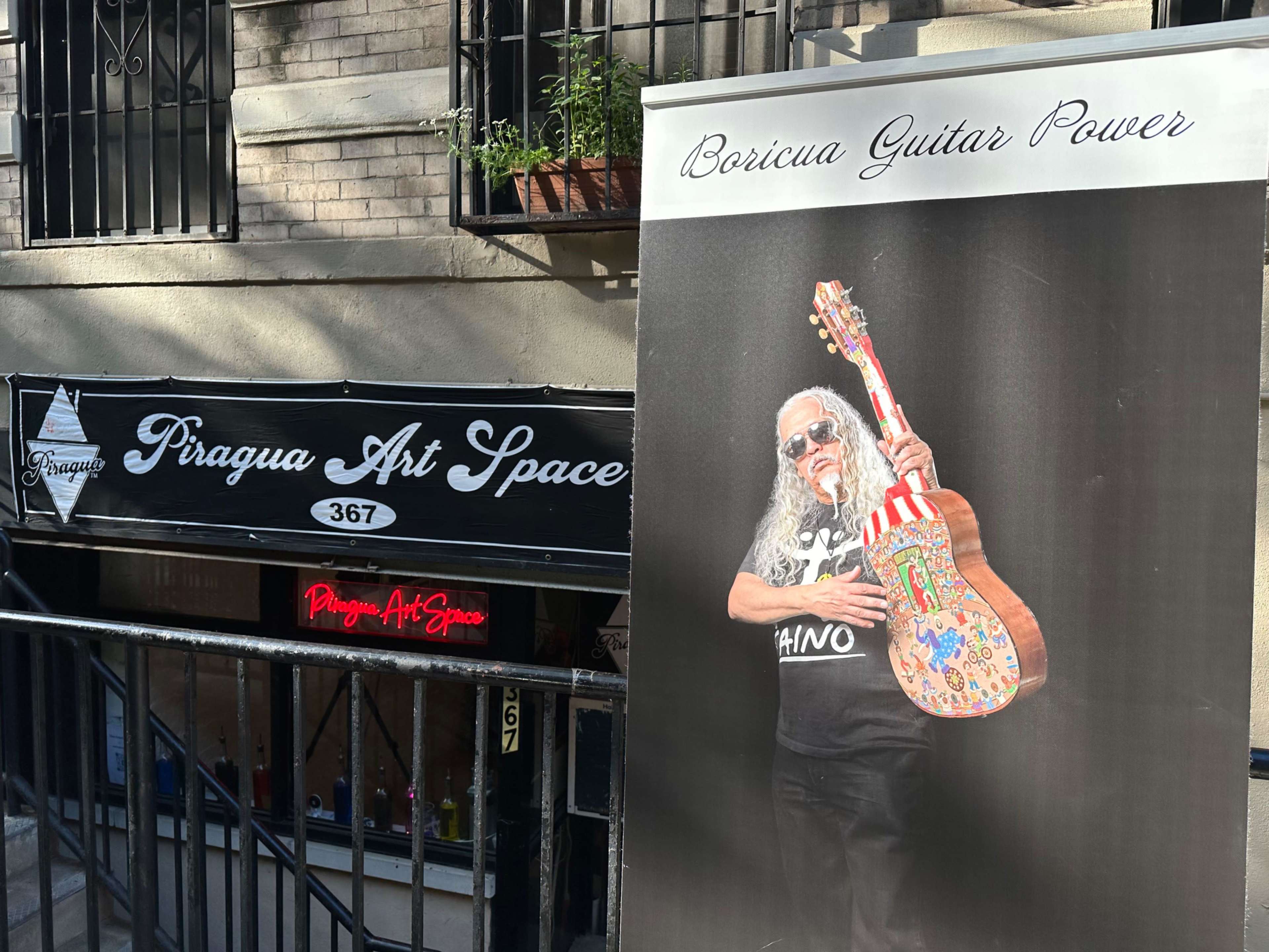 A street view of Piragua Art Space with a banner advertising "Boricua Guitar Power" featuring a man holding a decorated guitar.