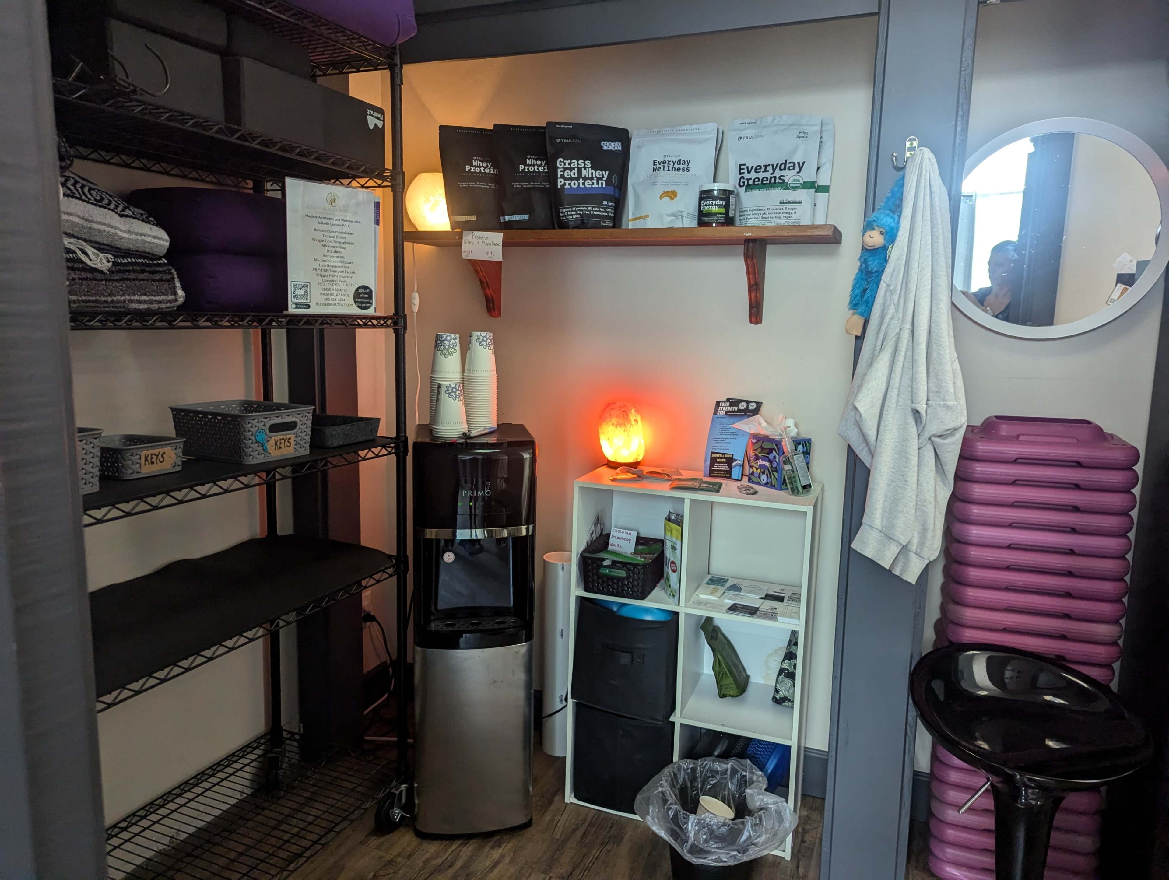 A small storage area with shelves containing bags, cups, a water dispenser, a trash can, and yoga mats neatly arranged on the floor.