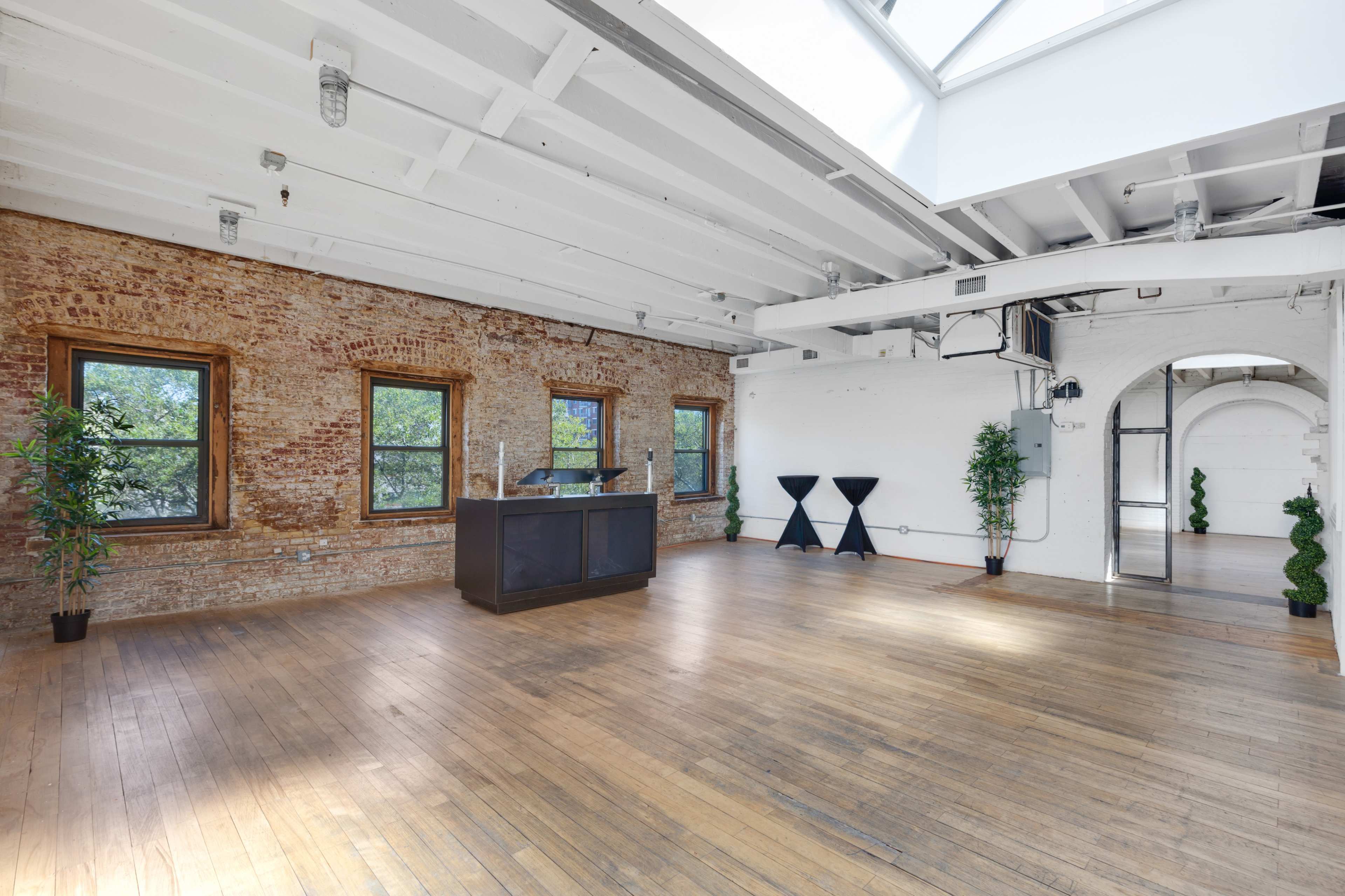 The image shows a spacious, empty room with exposed brick walls, large windows, and wooden flooring, featuring a reception desk and two black display stands.