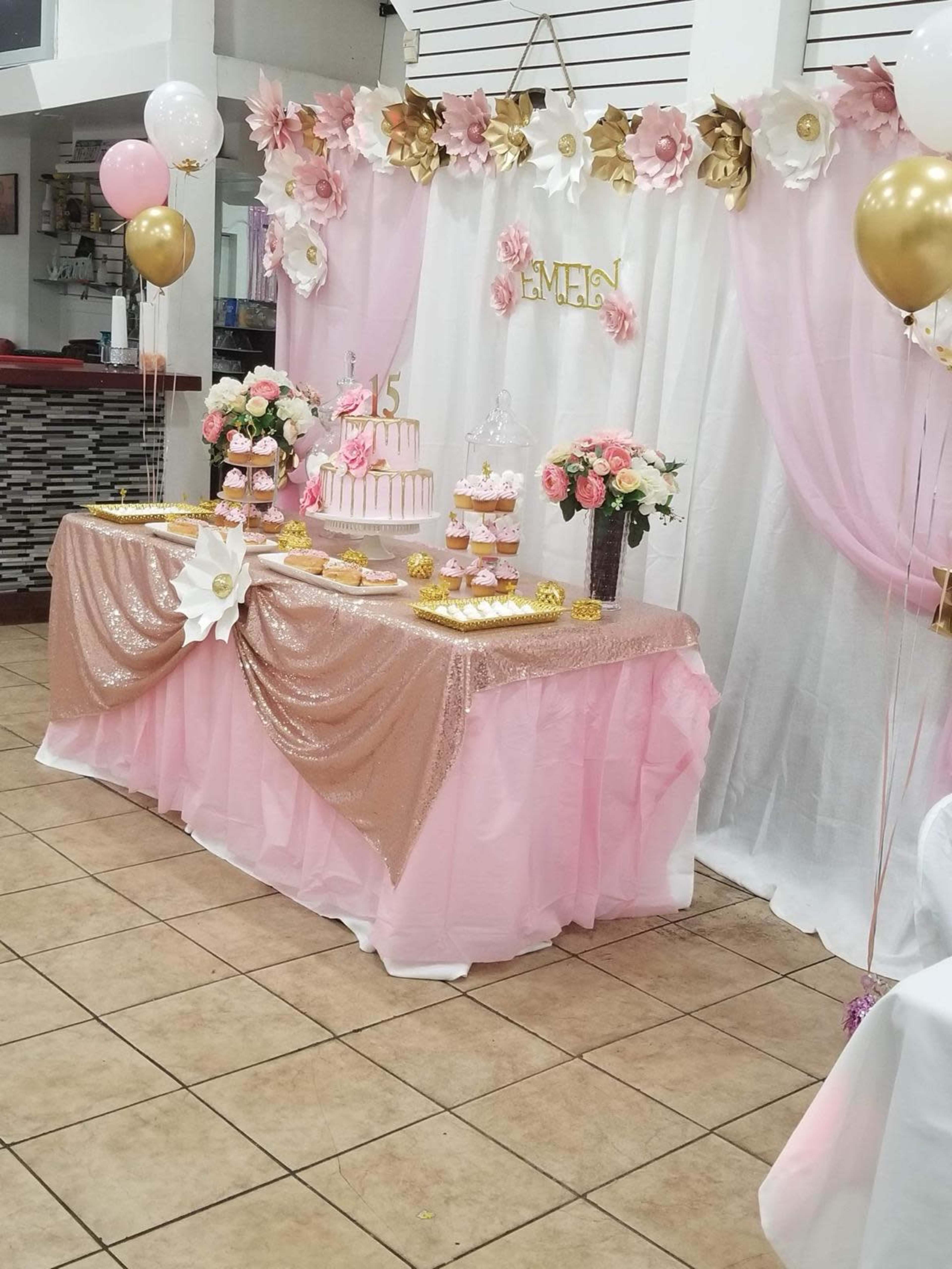 A decorated table with a pink and gold theme features a tiered cake, cupcakes, and floral arrangements against a backdrop of draped fabric and paper flowers.