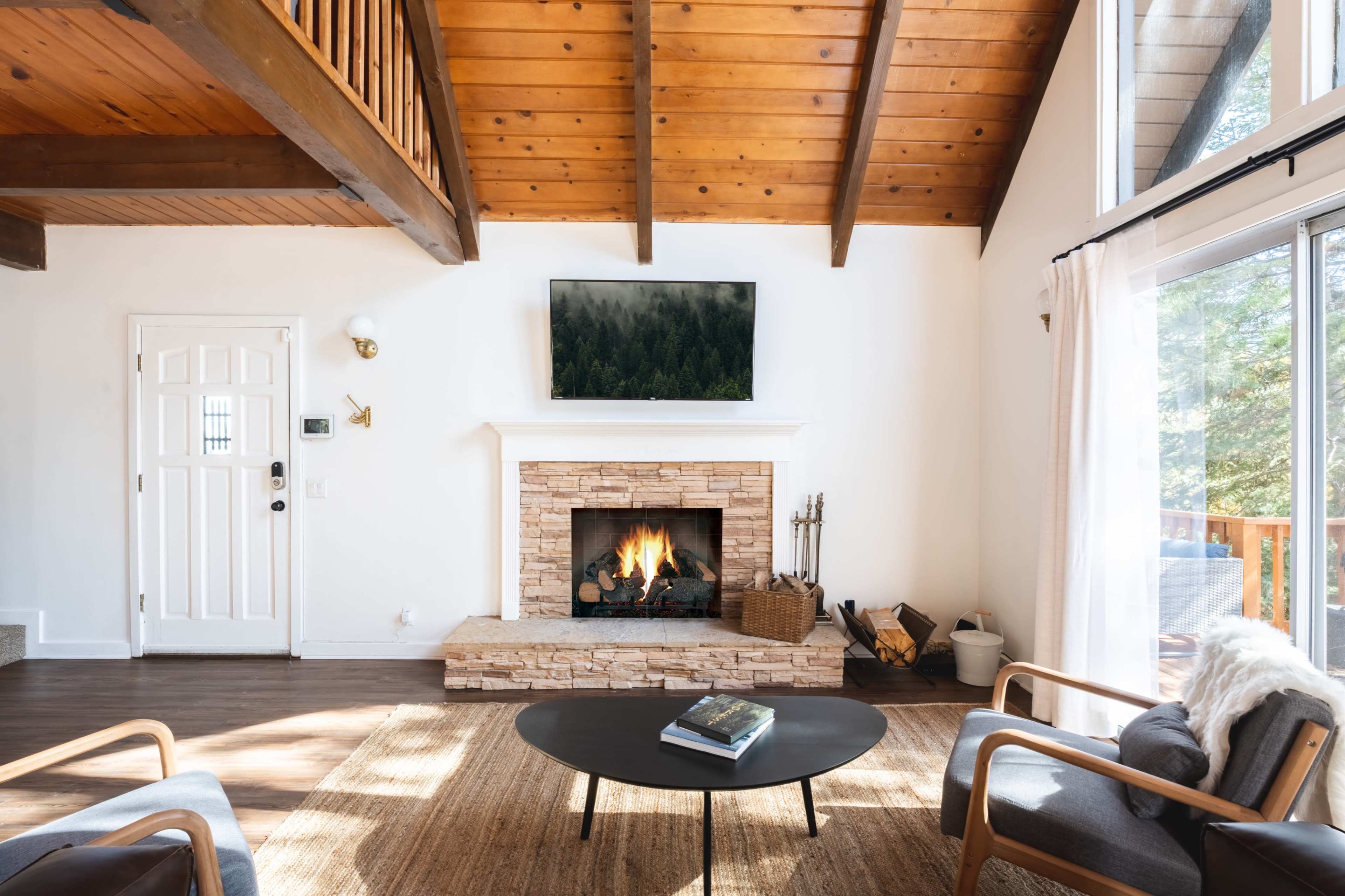 The image shows a cozy living room featuring a stone fireplace, a television mounted above it, wooden beams on the ceiling, and a coffee table in the center.