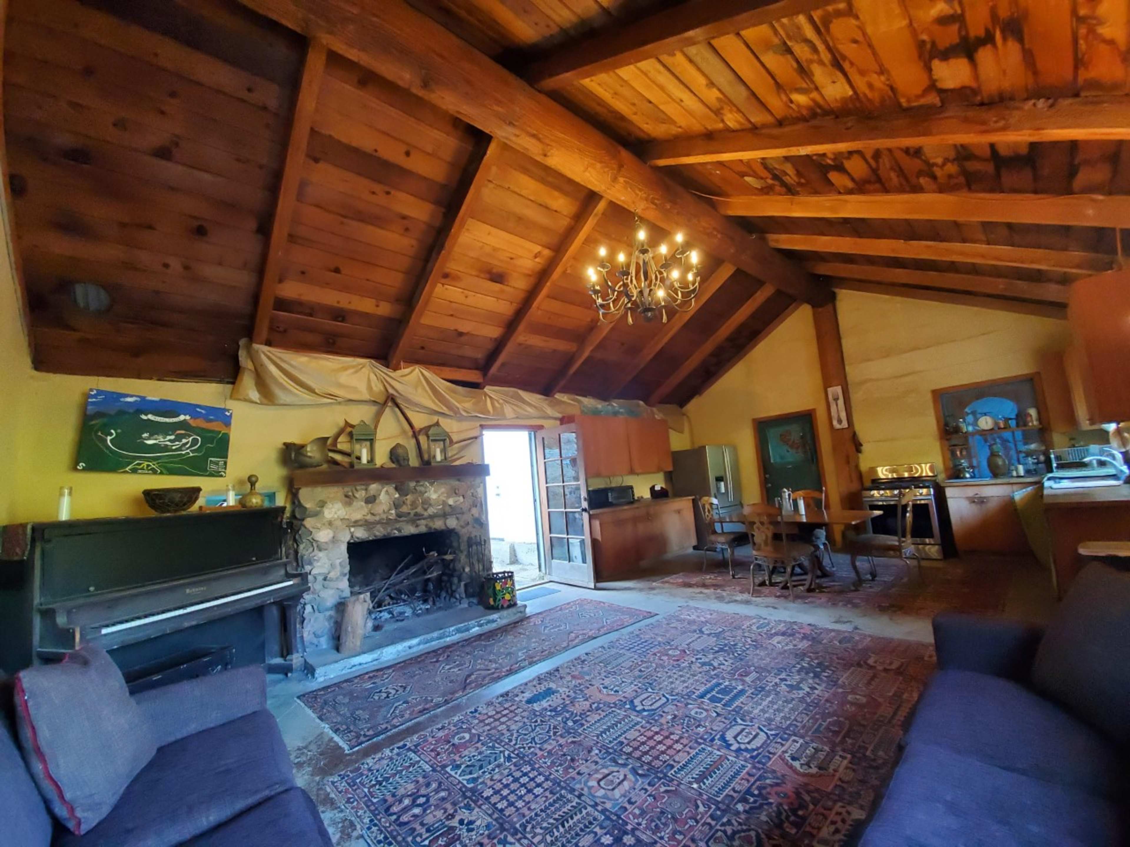 A rustic living space with wooden beams, a stone fireplace, a piano, and a dining area, all illuminated by natural light from an open door.